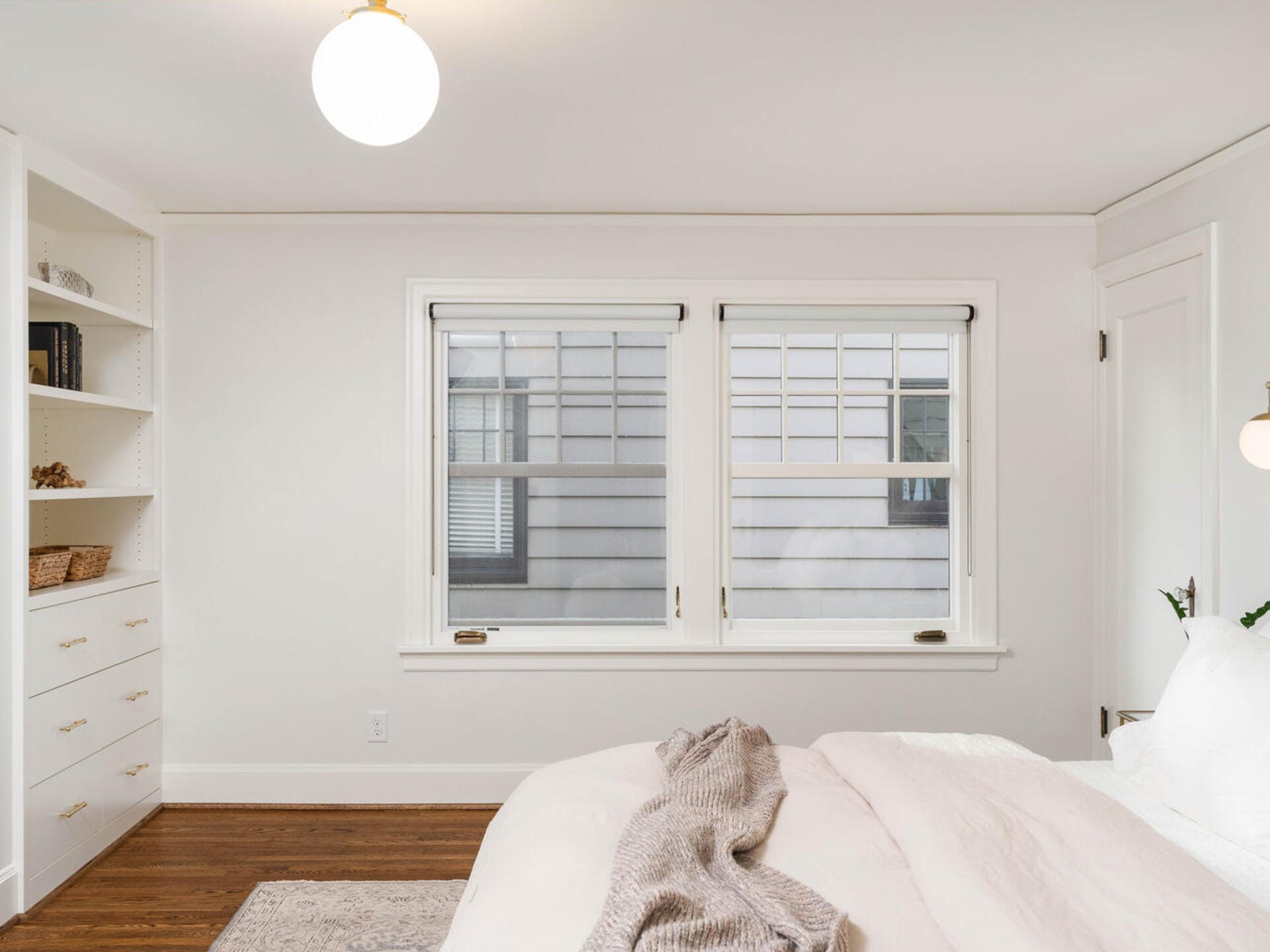 A bright, minimalist bedroom with white walls and bedding. Two windows on the far wall let in natural light. Built-in shelves on the left hold books and baskets. A cozy throw blanket is draped over the bed. A round ceiling light hangs above.
