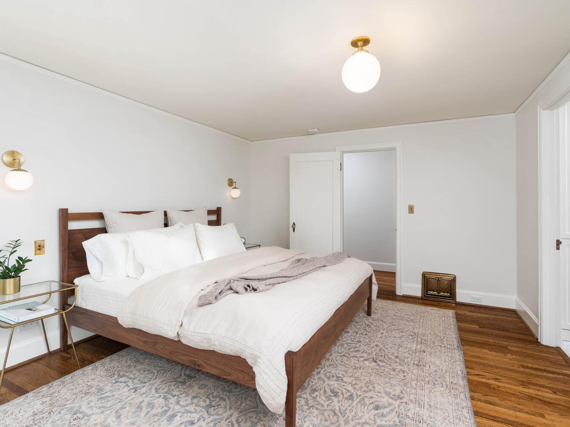 A modern bedroom features a wooden bed frame with white bedding, a soft gray blanket, and pillows. Two wall-mounted lamps and a ceiling light illuminate the space. The room includes a patterned rug, hardwood floor, small side table, and white walls.