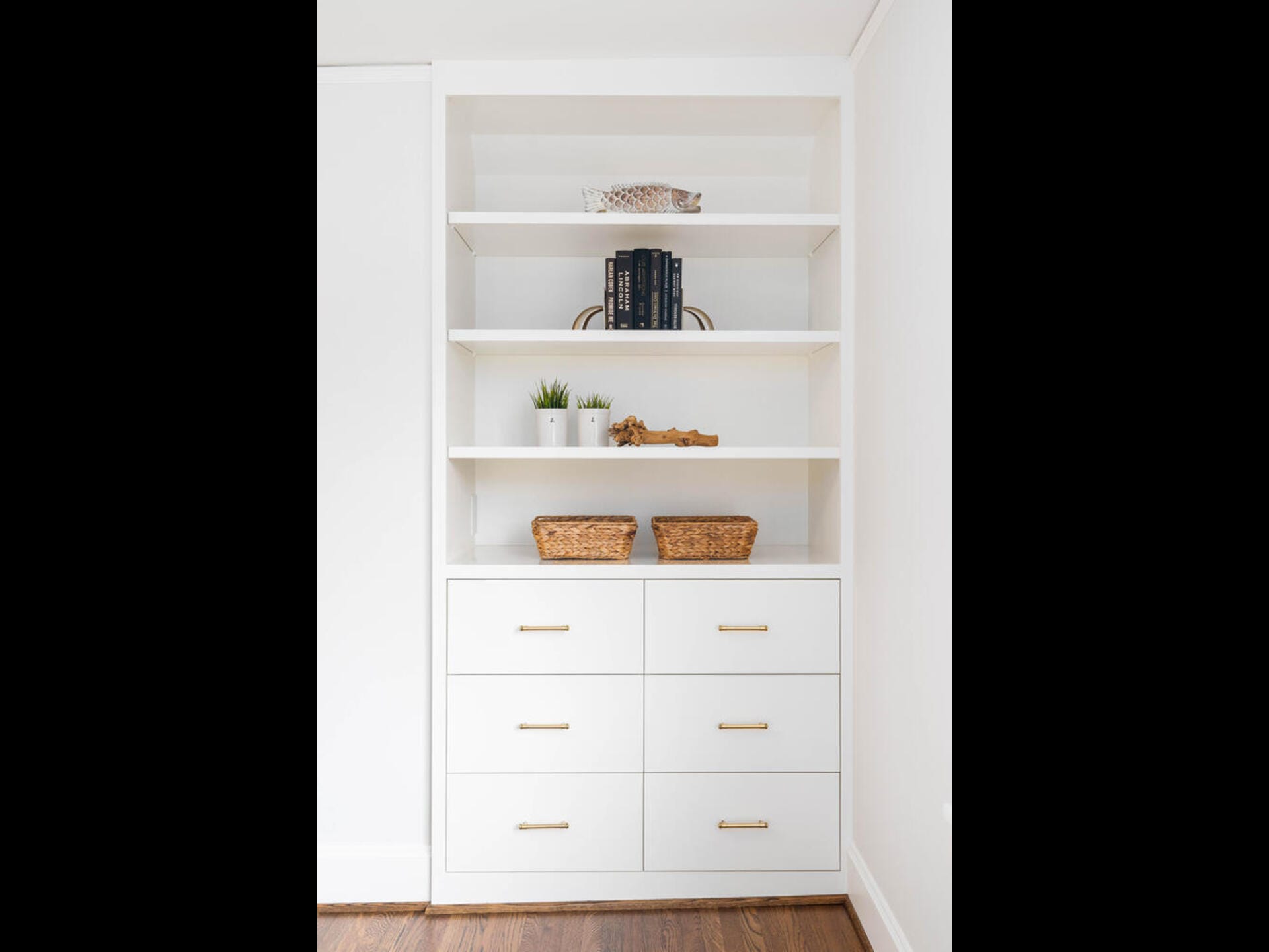 A white built-in shelving unit with four shelves and lower drawers. The shelves hold decor items such as small plants, books, and decorative objects. Two wicker baskets are placed side by side on one of the lower shelves. The floor is wooden.