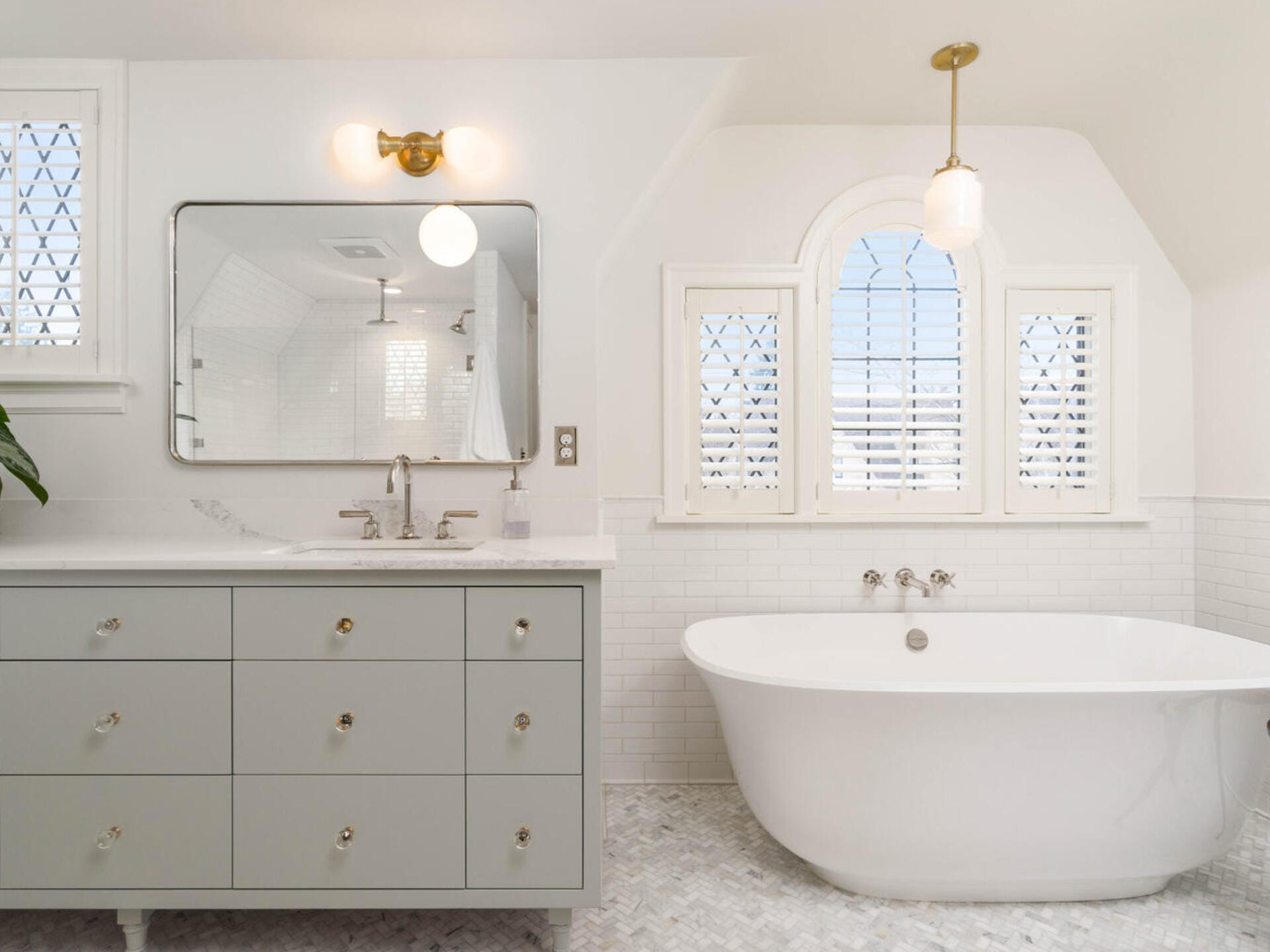 A bright bathroom with a freestanding bathtub, a large mirror above a light gray vanity with six drawers, and wall-mounted lighting. The room features a window with shutters, white subway tiles, and a patterned floor.