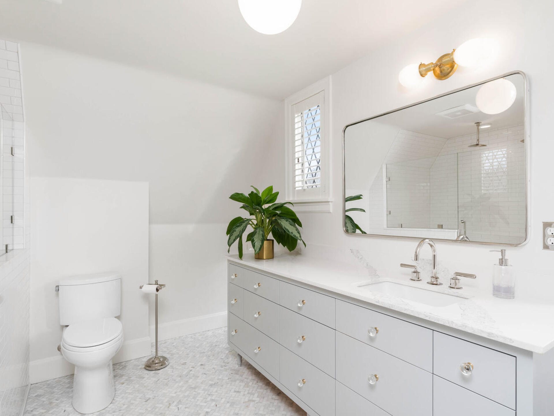 A bright, modern bathroom with a white and gray color scheme featuring a large mirror, double sink vanity, potted plant, toilet, and glass-enclosed shower. A circular ceiling light provides illumination.