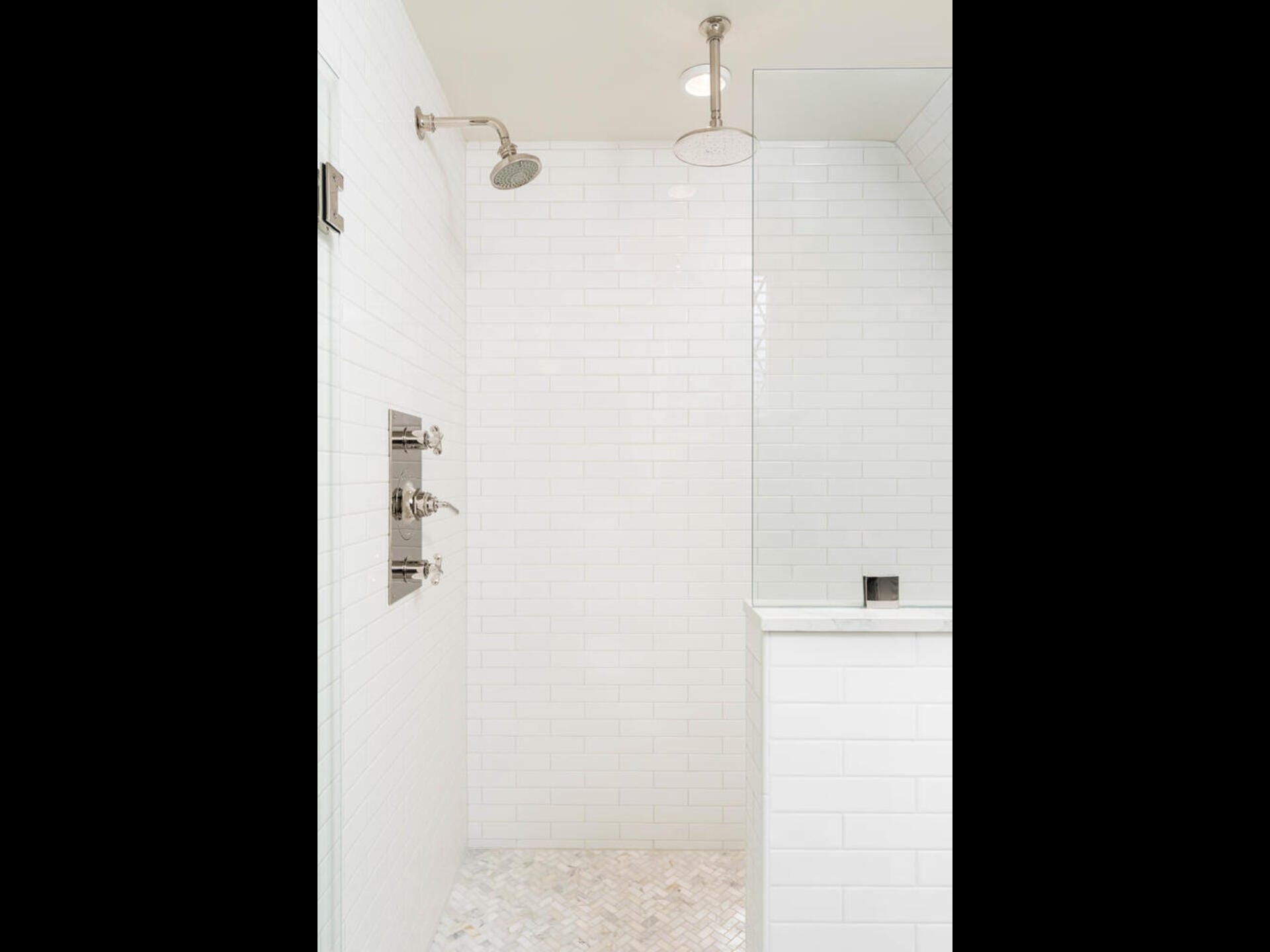 A modern, white-tiled shower with a rain showerhead and metal fixtures. The space features a glass partition and a tiled floor with a subtle herringbone pattern.