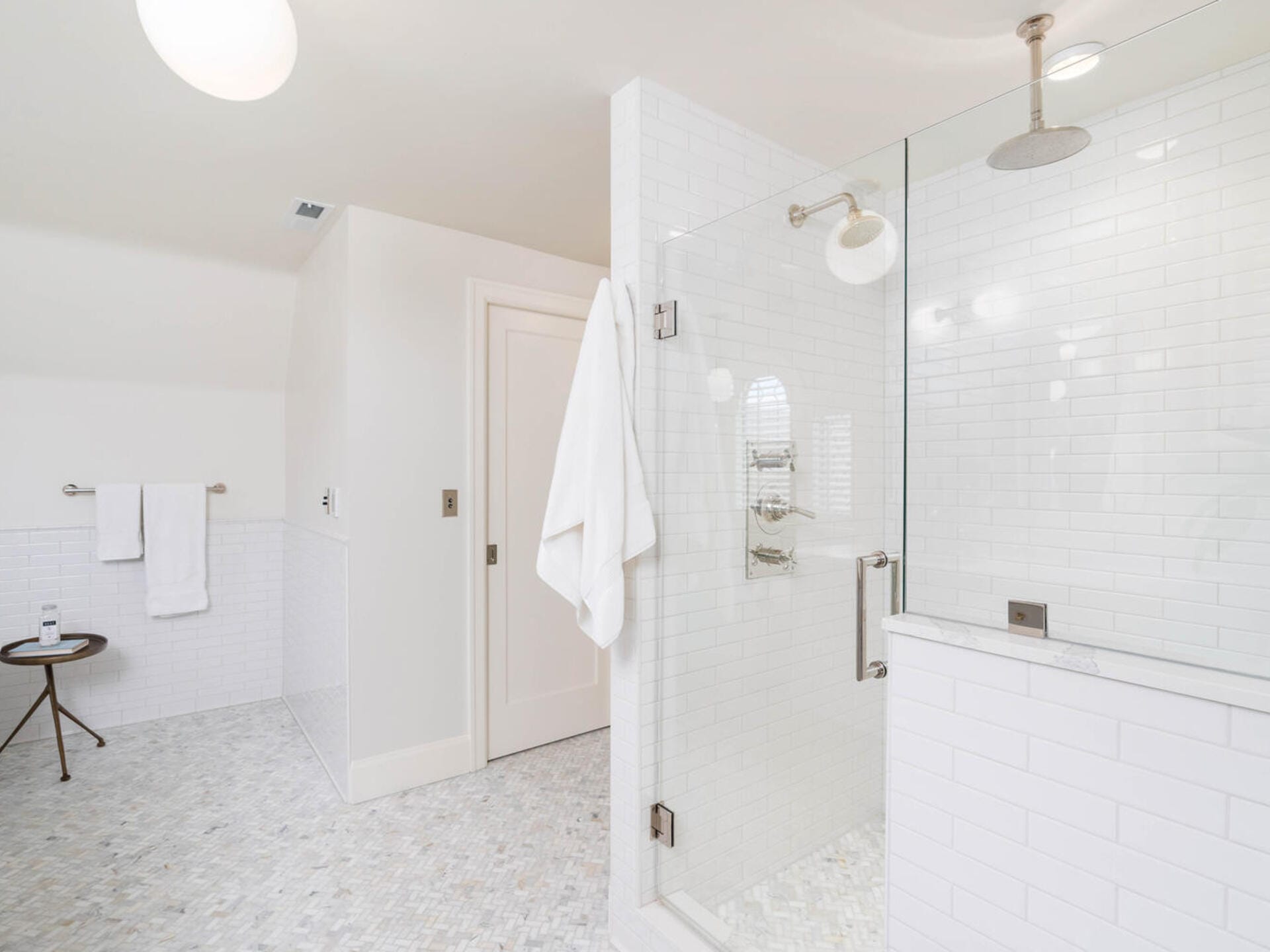 A modern bathroom with white subway tiles and a glass-enclosed shower. Towels hang near the shower and on a rack on the wall. Theres a small round table with a bottle on it. The floor is tiled with a light pattern, and the room is well-lit.