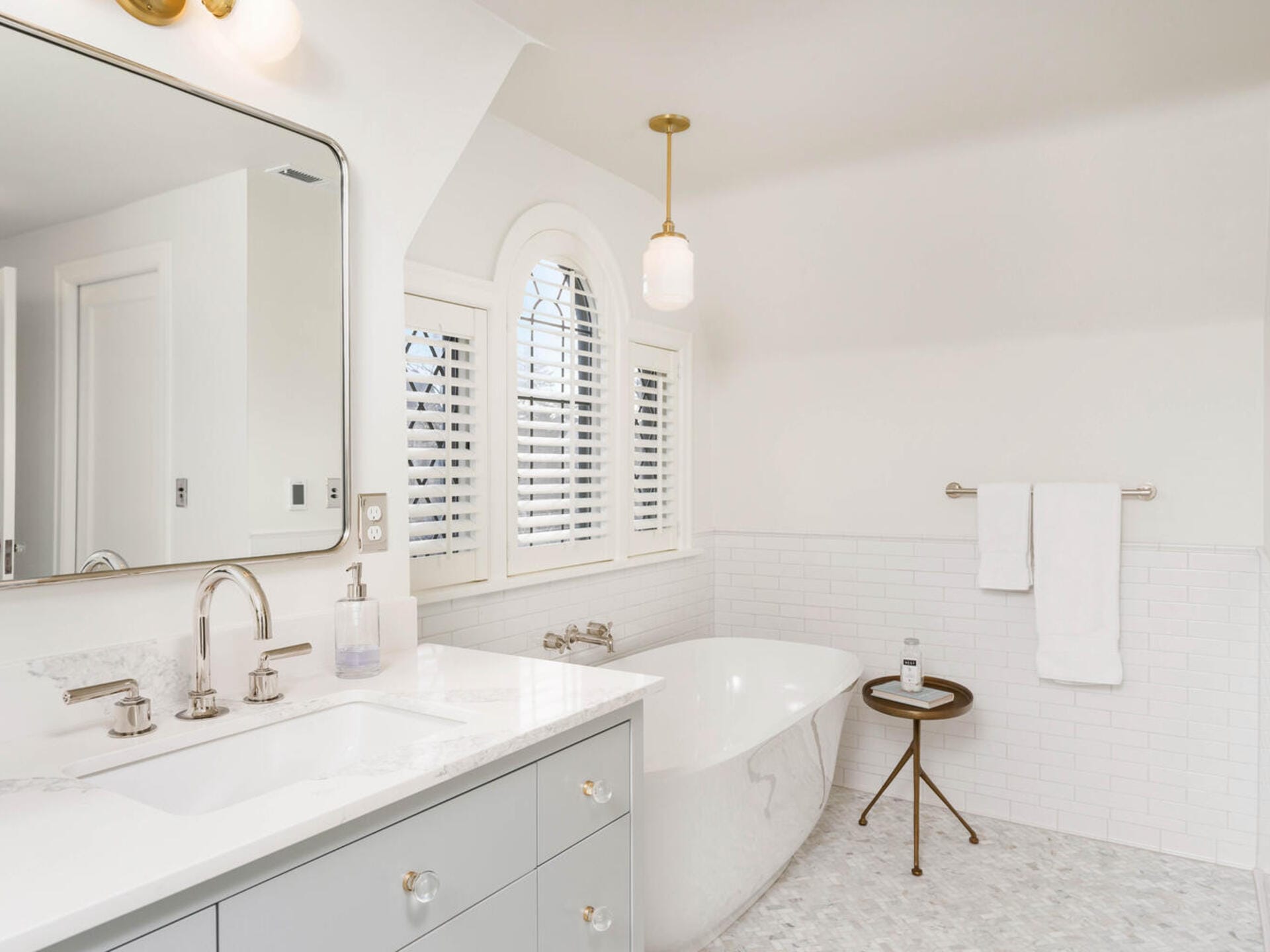 A bright, modern bathroom with a white freestanding bathtub, a curved window with shutters, a light grey vanity with a large mirror, and a small round table holding a candle. The walls and floor are tiled in white and light grey.