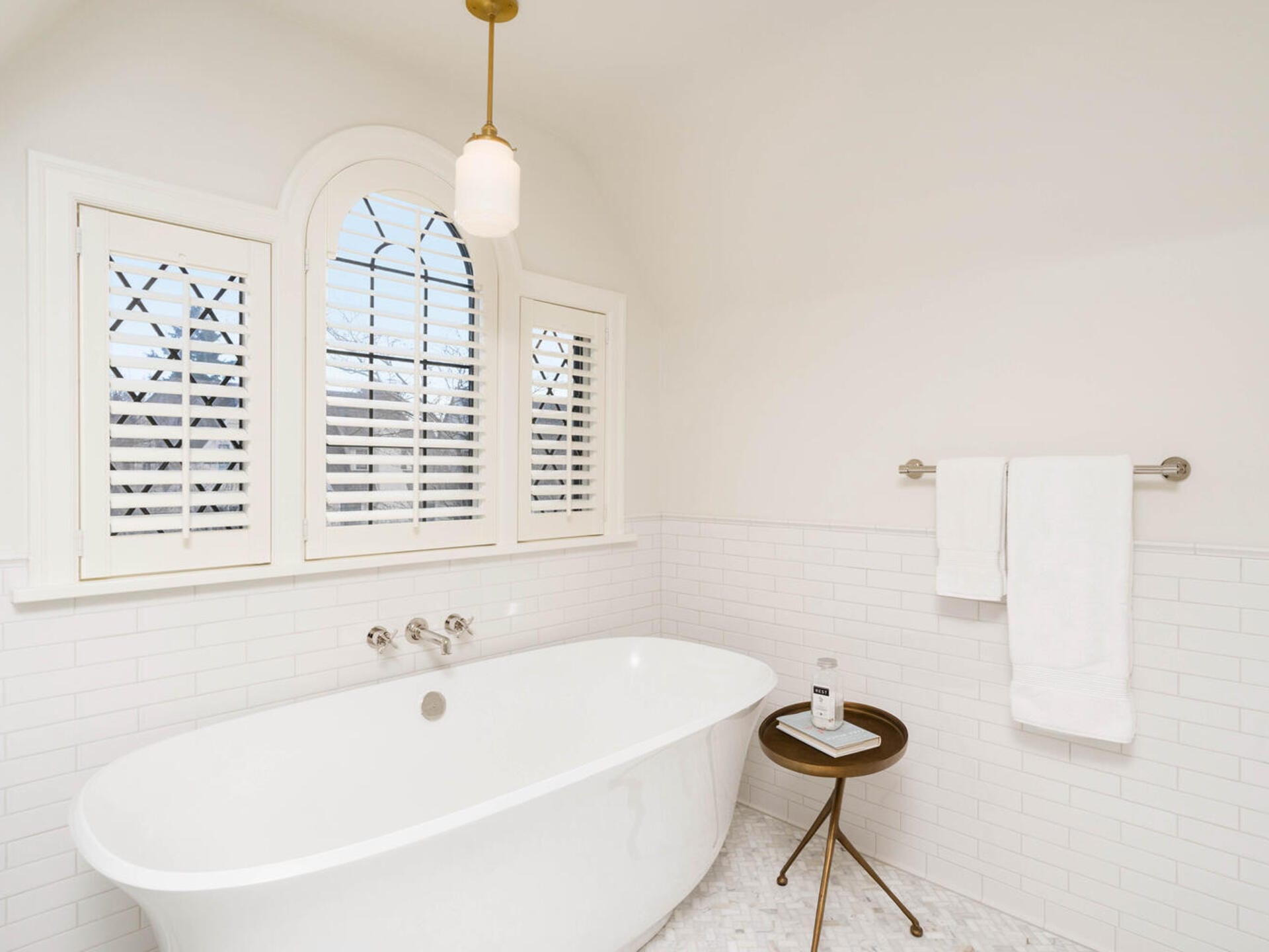 A serene bathroom with a white freestanding bathtub, arched window with shutters, and white subway tile walls. A small round table holds a candle and book. White towels hang on a rail nearby, under a pendant light.