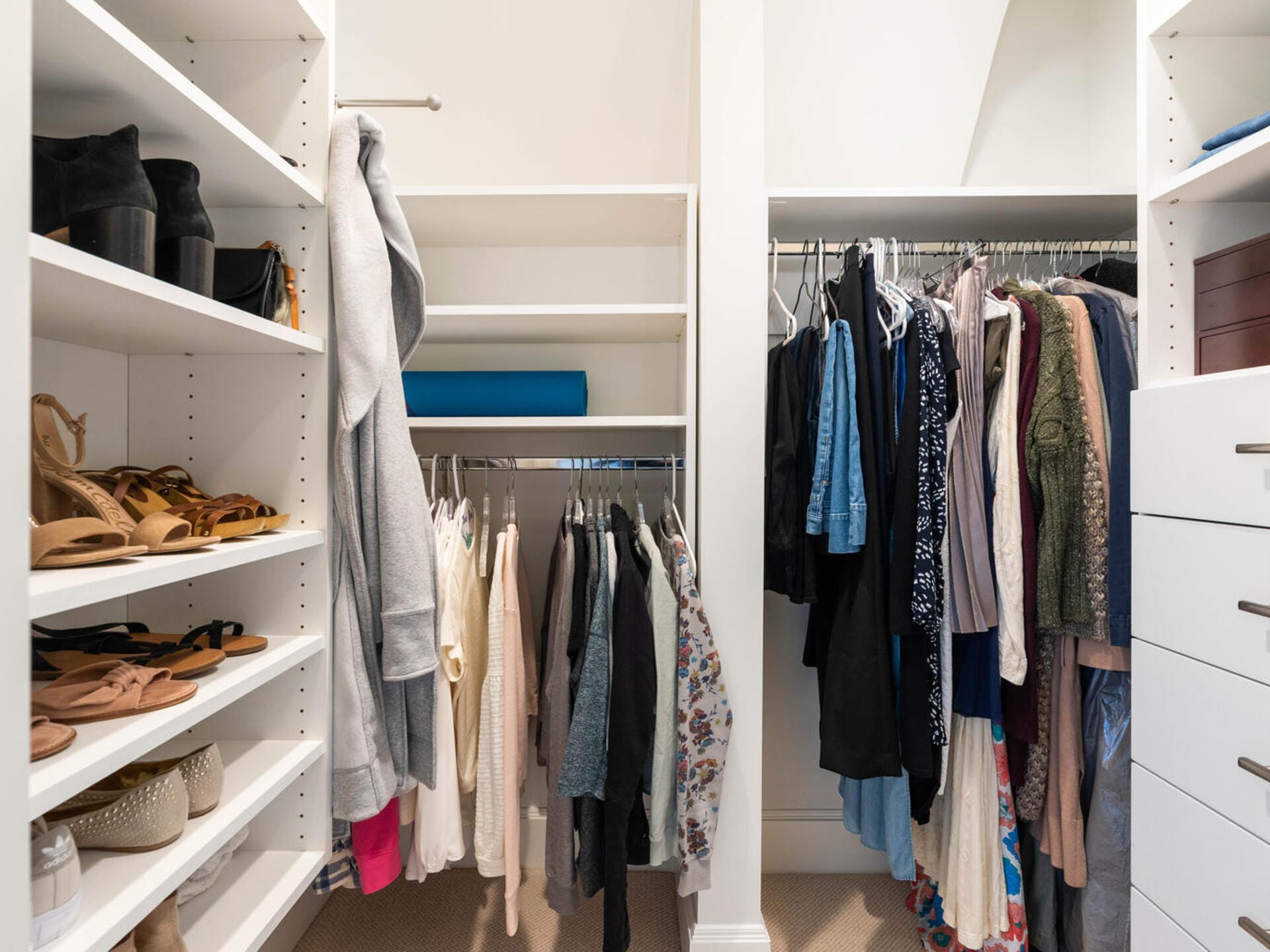 A neatly organized walk-in closet with various clothes hanging on rods. Shelves on the left hold shoes and folded clothes. On the right, drawers and more hanging clothes are visible. The space is well-lit and spacious.
