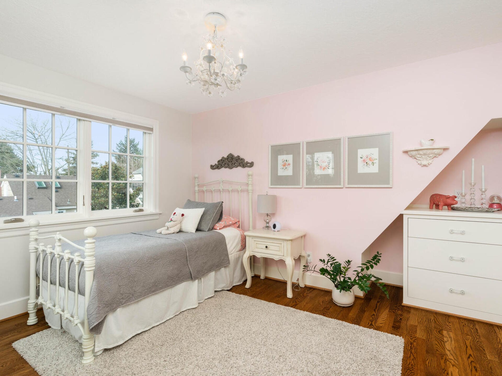 A cozy bedroom with a single bed featuring gray and pink bedding. The walls are light pink with framed floral artwork. A white dresser and chandelier add a touch of elegance. A window lets in natural light, and theres a potted plant on the hardwood floor.