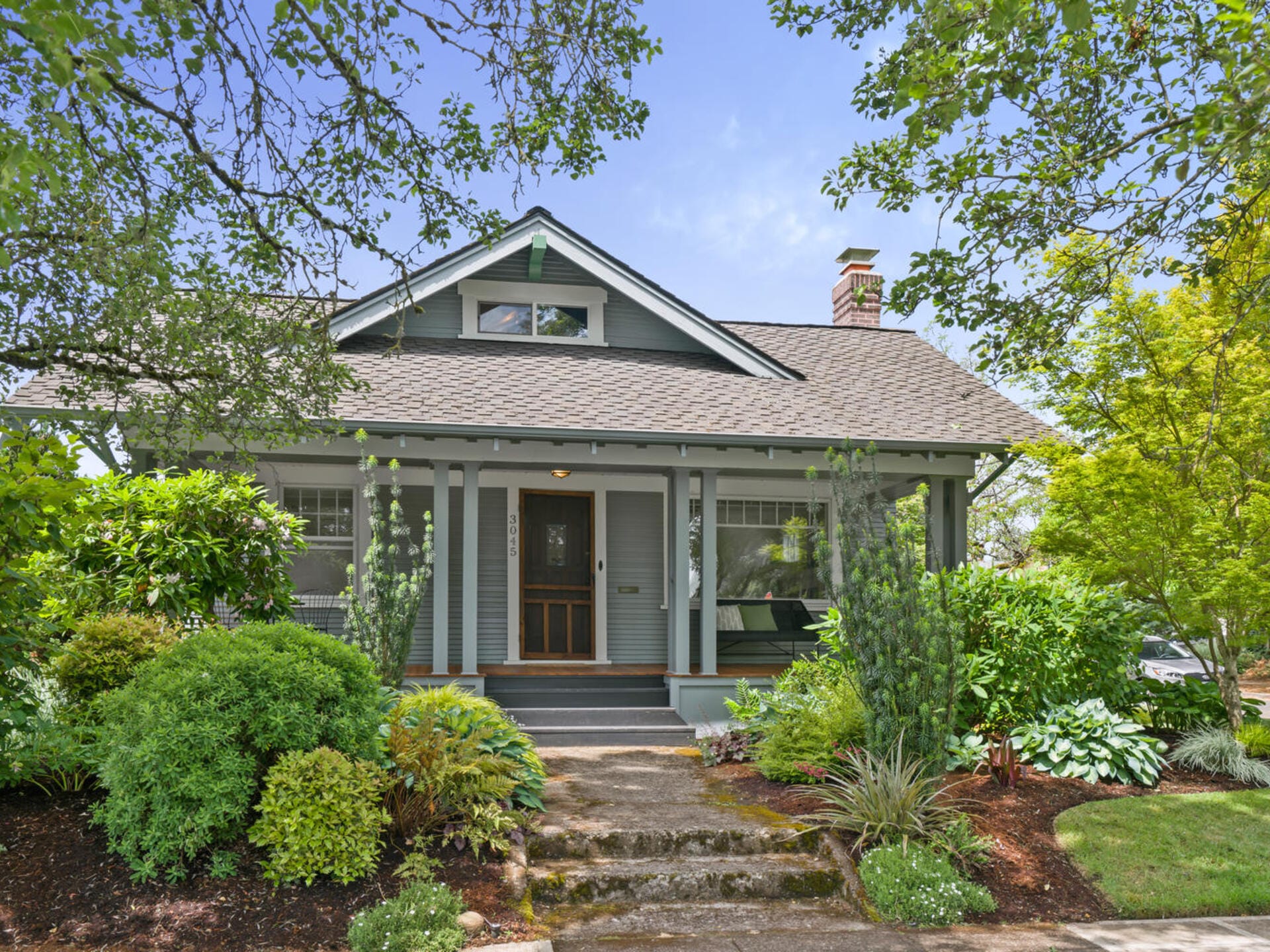 A charming, light blue house with a gabled roof and front porch. Surrounded by lush greenery and trees, it features a welcoming entrance with steps leading up. A bright, clear sky is visible in the background.