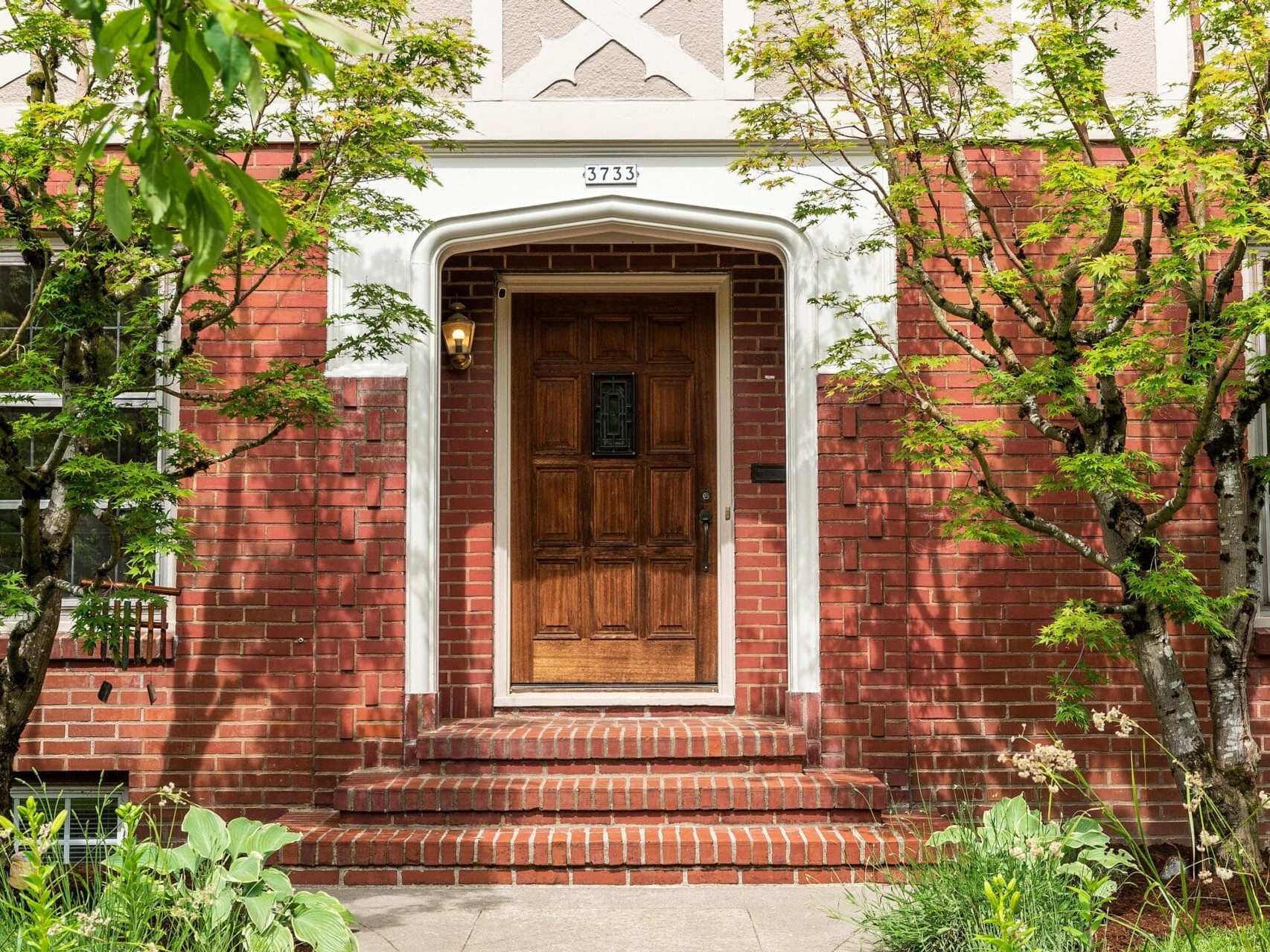 A wooden door with decorative glass pane set in a brick facade. Two small trees frame the entrance, and the house number 3733 is displayed above. Shadows from the trees are cast on the brick steps leading to the door.