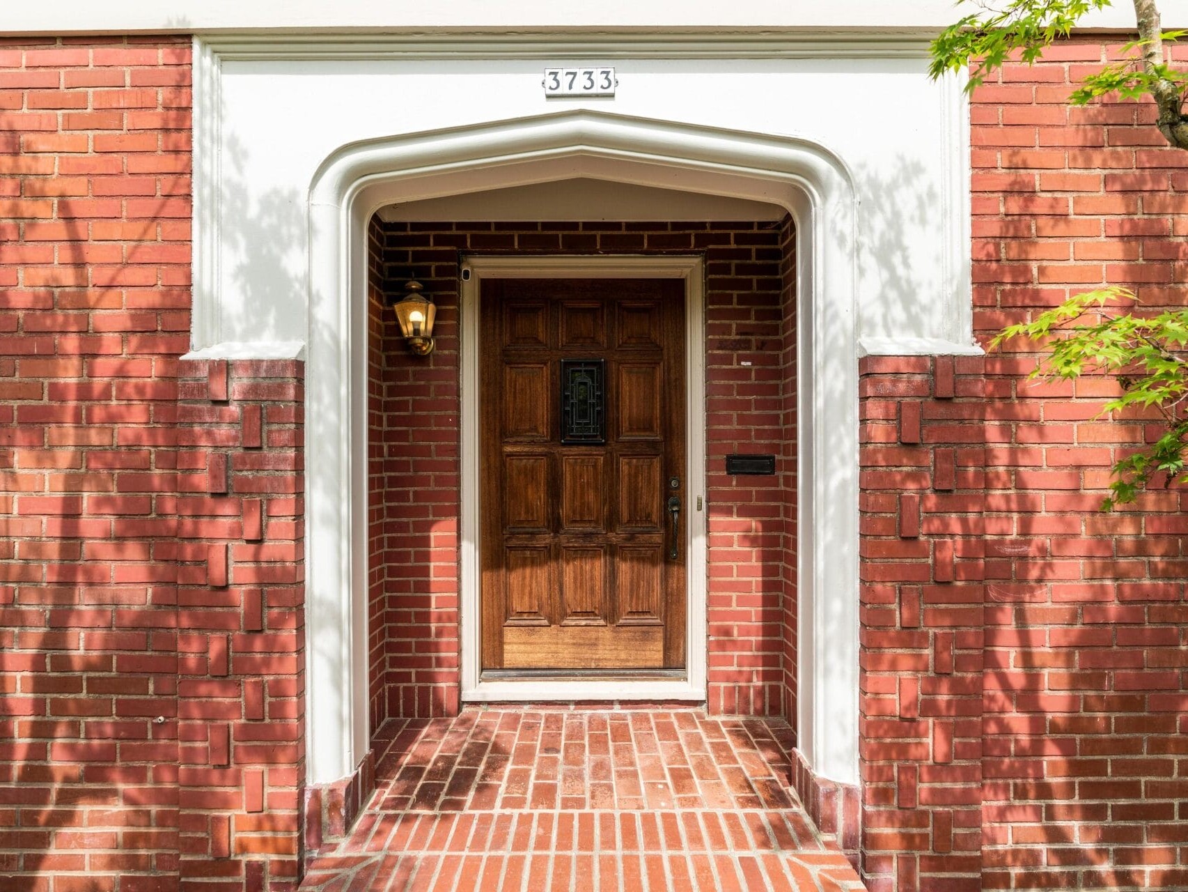 A brick front entrance of a building with the number 3733 above a wooden door. A small lantern-style light is on the wall to the left of the door, and a leafless tree casts shadows on the bricks.