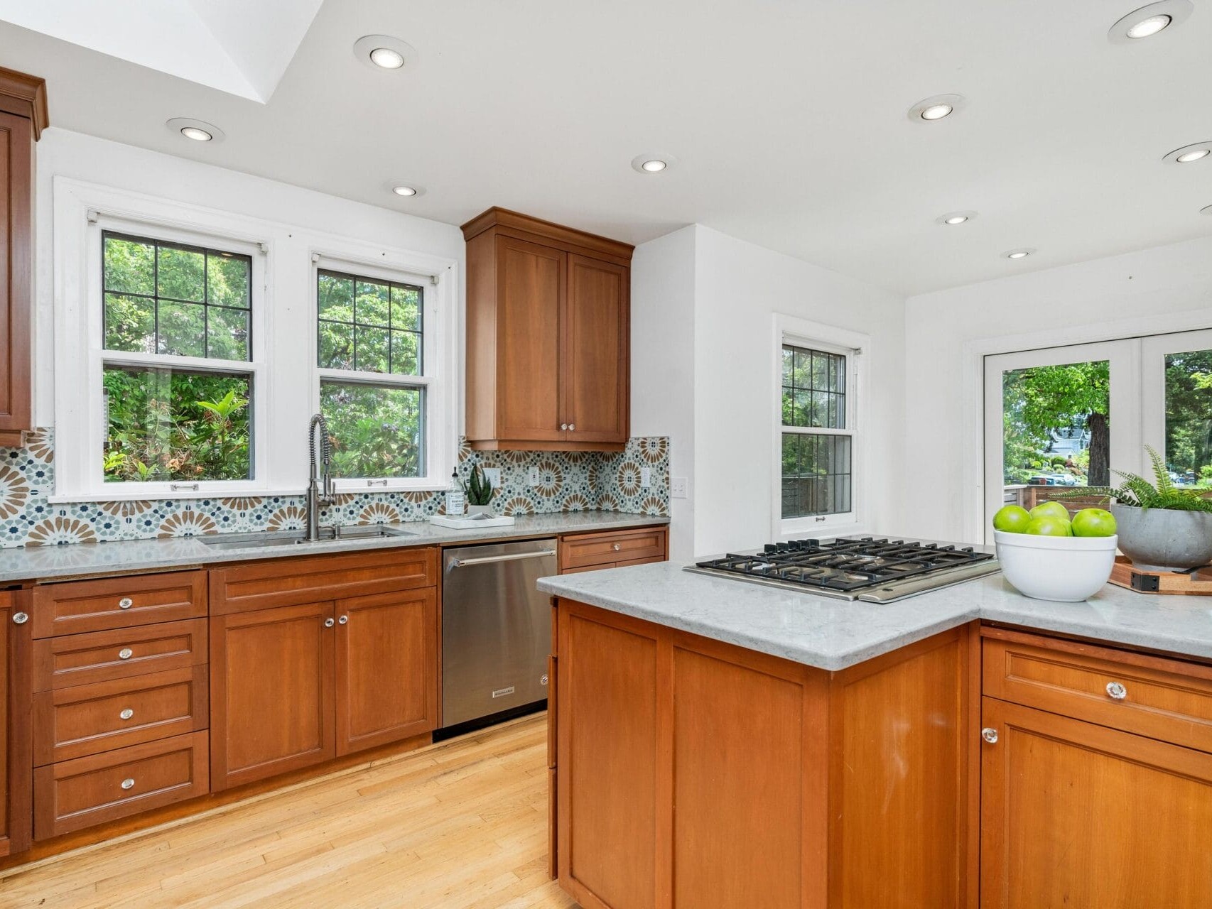 A bright kitchen with wooden cabinets and a decorative tile backsplash. It features a stainless steel dishwasher, a stove on the island with a bowl of green apples, and large windows allowing ample natural light.