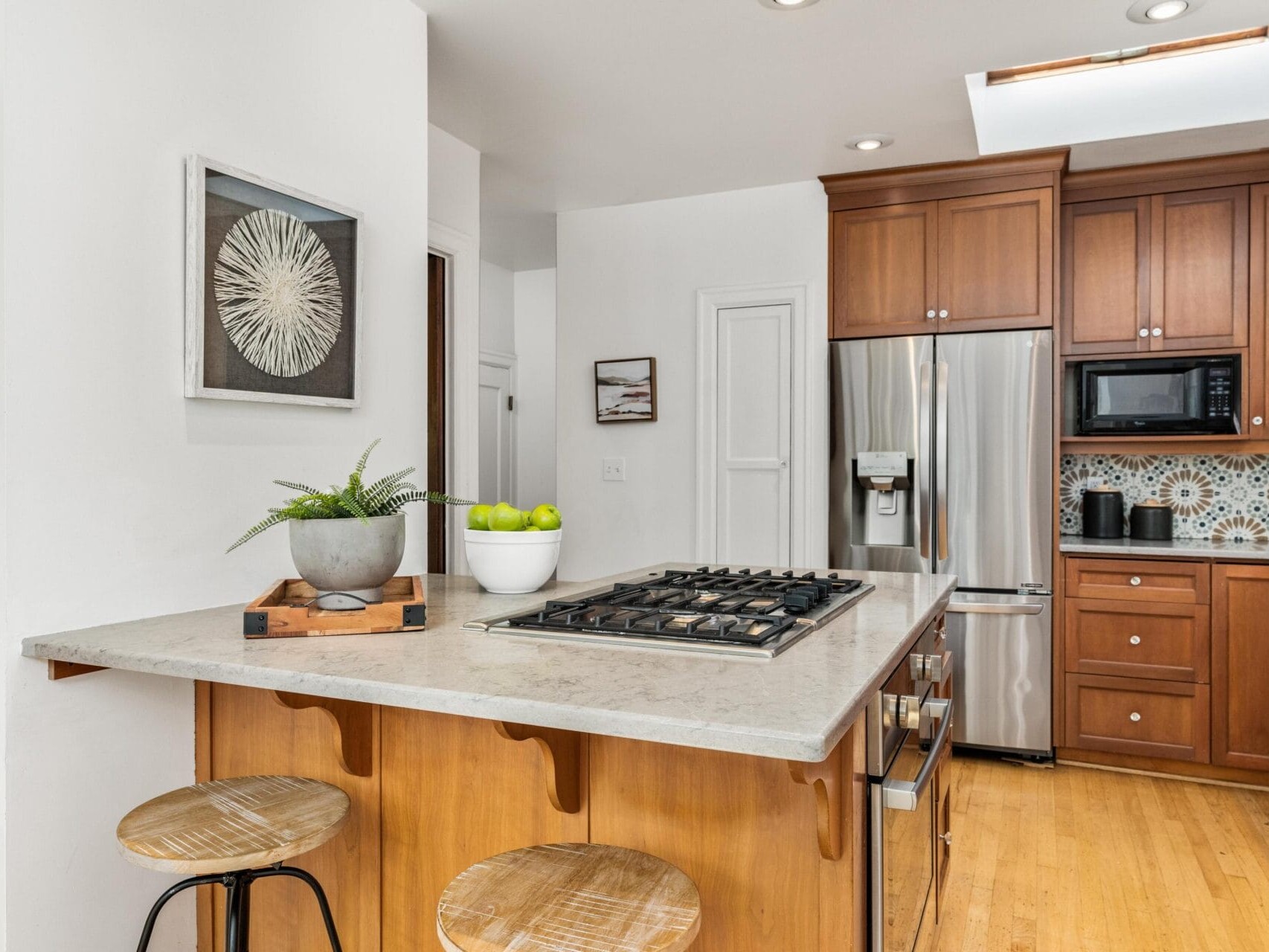 Modern kitchen with wooden cabinets and light wood flooring. Central island features a gas stovetop, two wooden stools, and a decorative plant. Stainless steel appliances, including a refrigerator and microwave, are built into the cabinetry. Wall art adds decor.