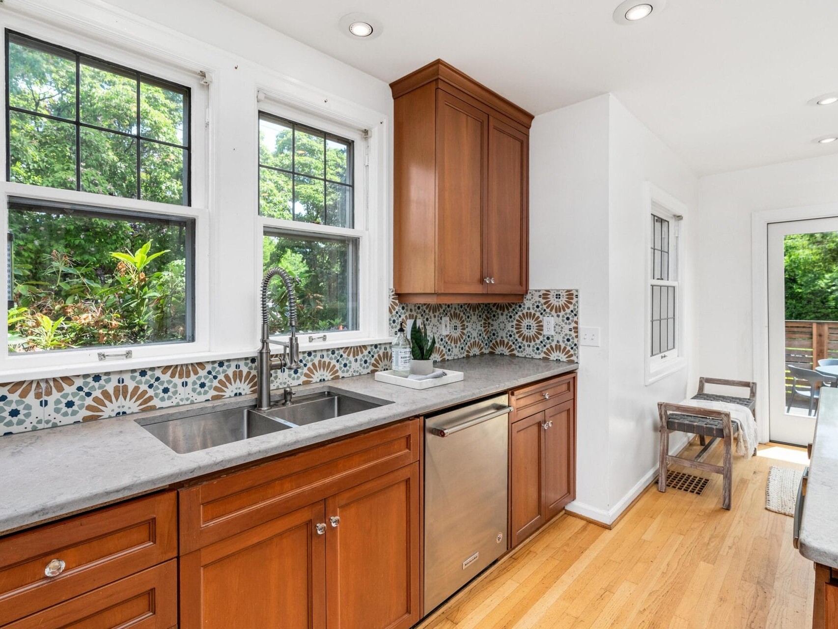 A bright kitchen with wooden cabinets, marble countertops, and a floral-patterned backsplash. Double sinks are under two large windows. A dishwasher is below the counter, and a door leads to a deck outside.