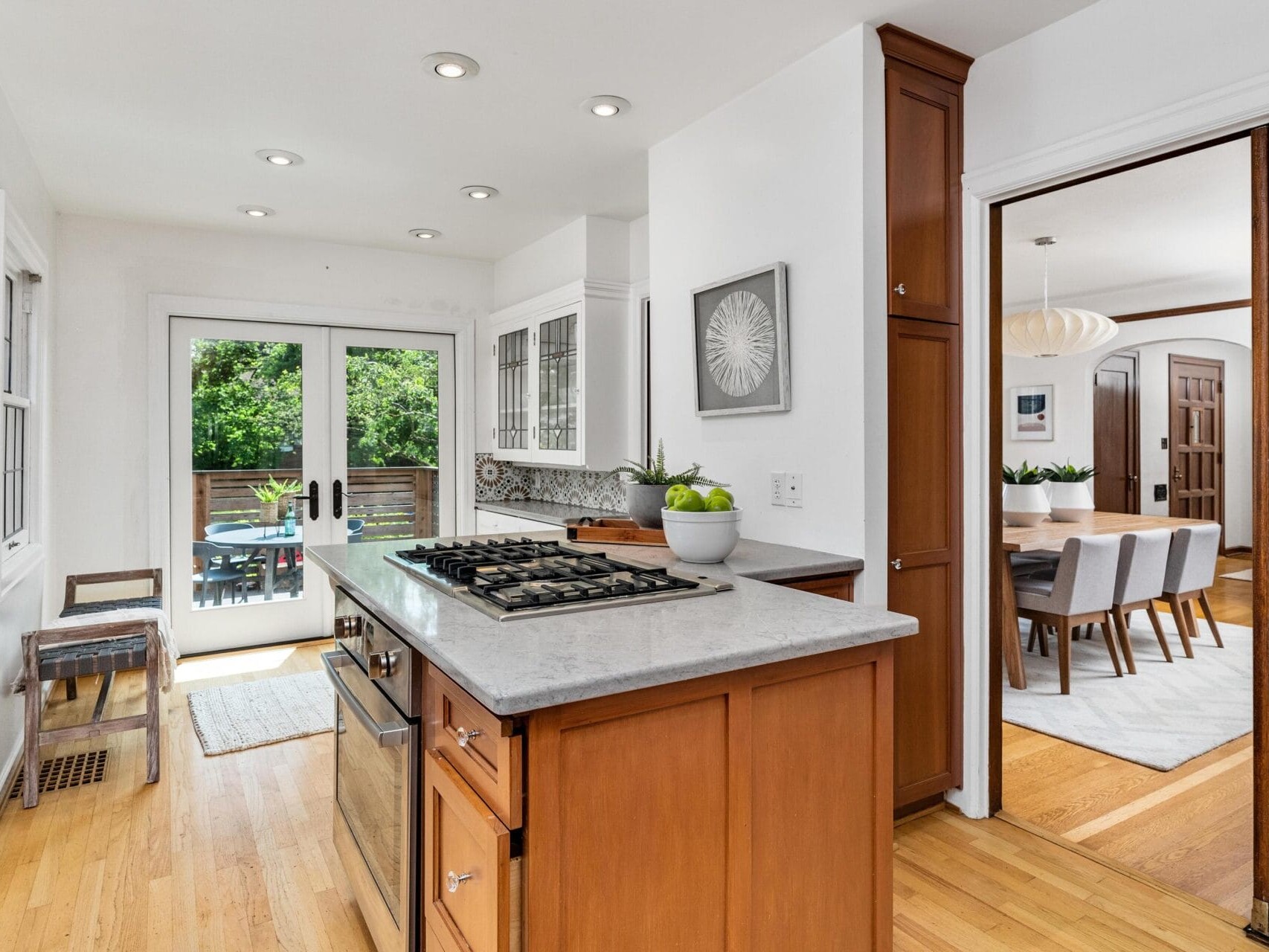 A modern kitchen with wooden cabinets, an island featuring a gas cooktop, and a light marble countertop. The kitchen opens to a dining area with a large table and chairs. Glass doors lead to a sunny outdoor patio. Bright and airy space with wooden floors.