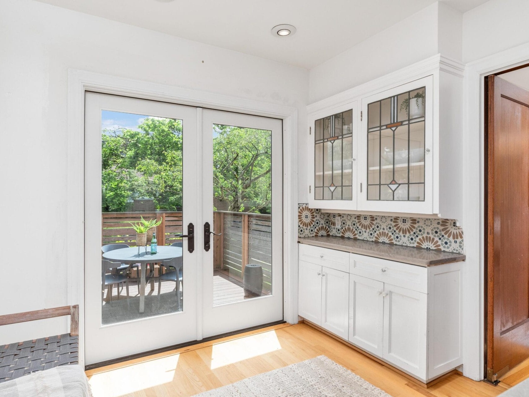 A bright room with a wooden floor, featuring white cabinets and a decorative backsplash. Glass doors lead to a patio with a table and chairs. Trees and greenery are visible outside, enhancing the fresh and inviting atmosphere.