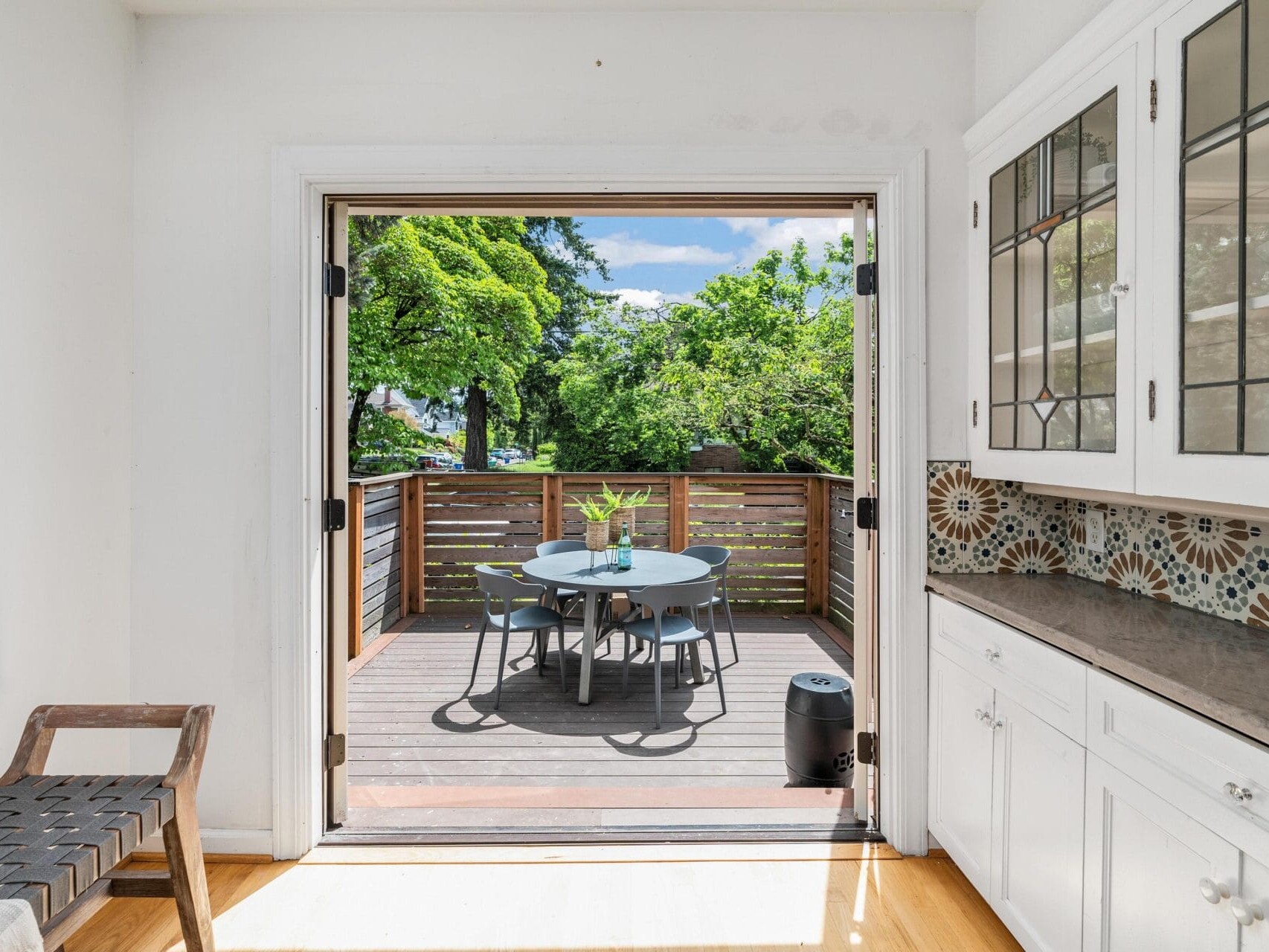 A bright kitchen leads to an outdoor deck through large open doors. The deck features a round table with chairs, surrounded by lush green trees. The kitchen has white cabinetry, decorative stained glass, and a mosaic tile backsplash.