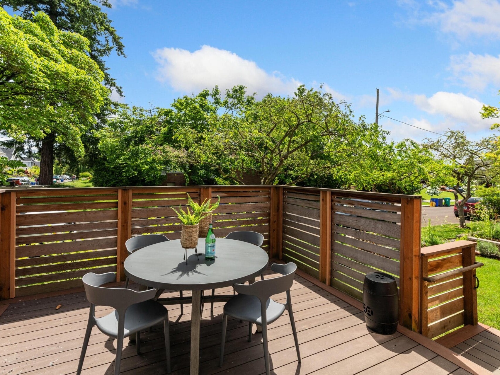 A wooden deck with a round table surrounded by four gray chairs. A potted plant and two bottles are on the table. The deck is bordered by a wooden fence with a lush, green yard and trees visible in the background under a blue sky.