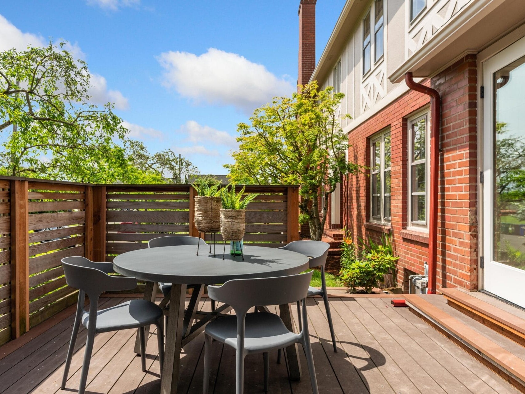 Outdoor patio with a round, black table and four chairs on a wooden deck. Two potted plants are on the table. The deck is bordered by a wooden fence and attached to a brick house. Green trees and a clear blue sky fill the background.