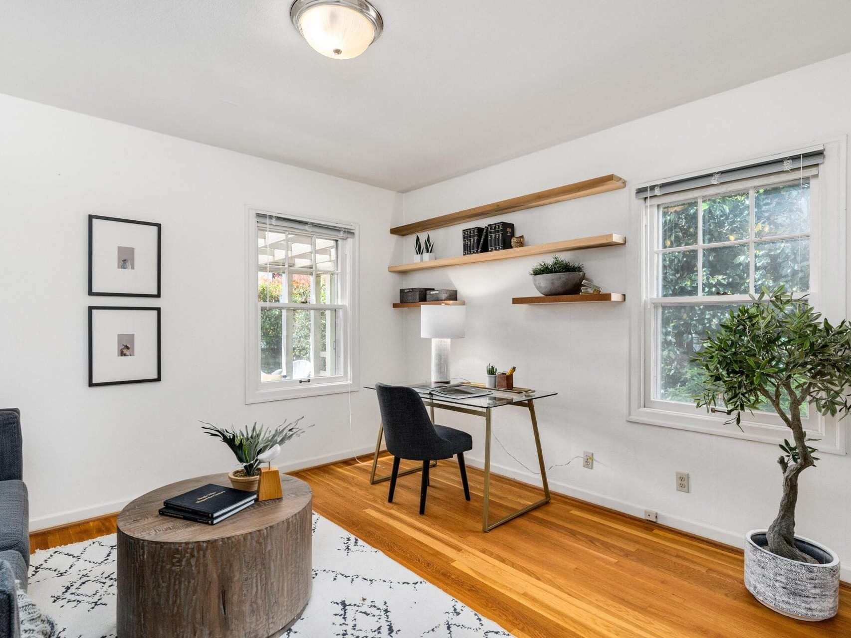 A bright home office with a minimalist design. It features a wooden desk with a lamp, a black chair, and floating shelves with books and decor. Theres a circular wooden coffee table, a rug, and a potted plant on a wooden floor, with two large windows.