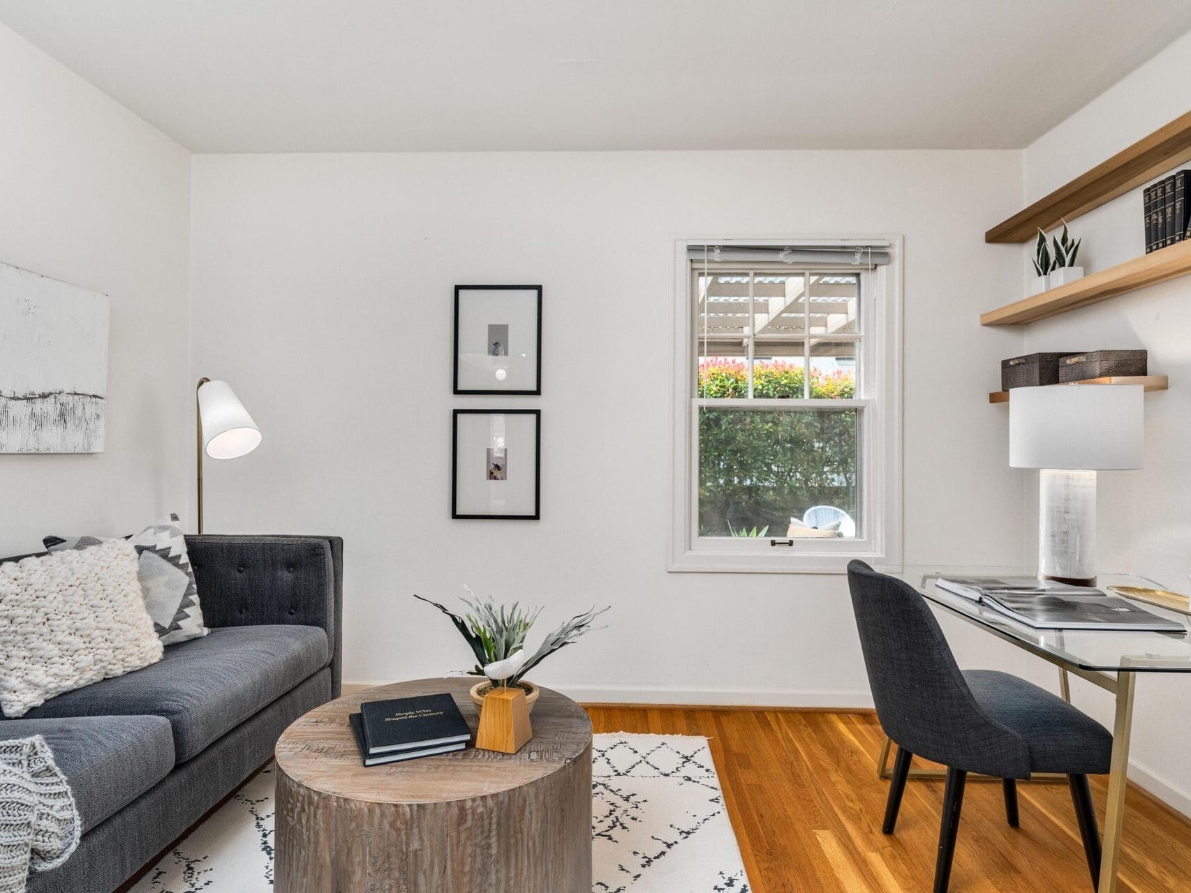 A cozy living room with a dark gray sofa adorned with pillows, a wooden coffee table, and a white floor lamp. A modern desk with a chair and lamp sits by the window, and two framed pictures hang on the wall above the sofa. Hardwood floors complete the space.