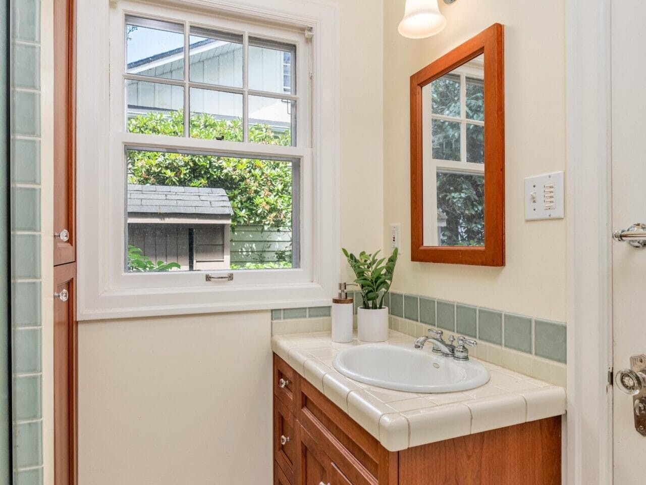 A small bathroom features a wooden vanity with a white countertop and sink. A wall-mounted mirror is above the sink, next to a window with an outdoor view. The room has tiled floors and light-colored walls, with a potted plant on the counter.