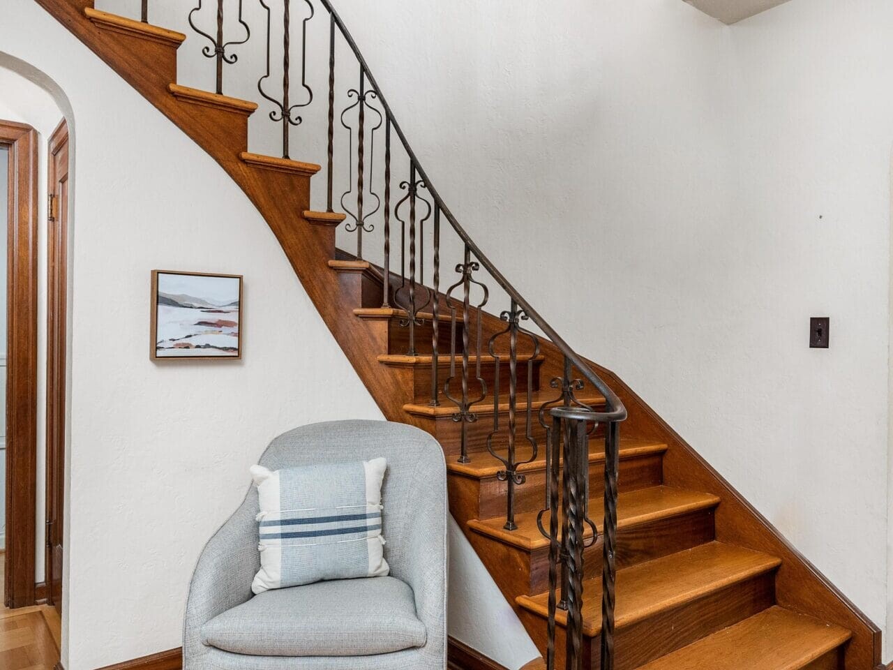 A cozy room featuring a wooden staircase with ornate iron railing, a modern gray armchair with a striped pillow, and a small framed picture on the wall. A stylish overhead light fixture adds elegance to the space.