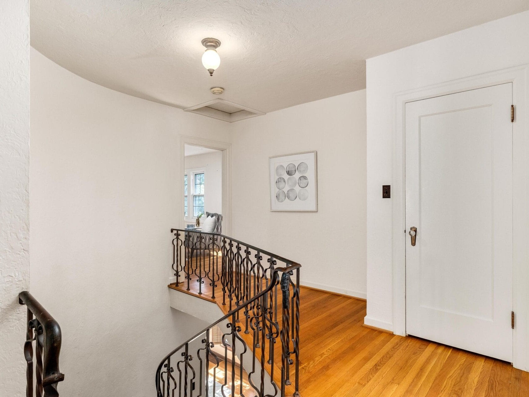 A hallway with wooden floors and white walls features a decorative black metal railing along the staircase. A white door is on the right, and a framed picture with circular patterns is on the wall. Natural light filters in from an adjoining room.