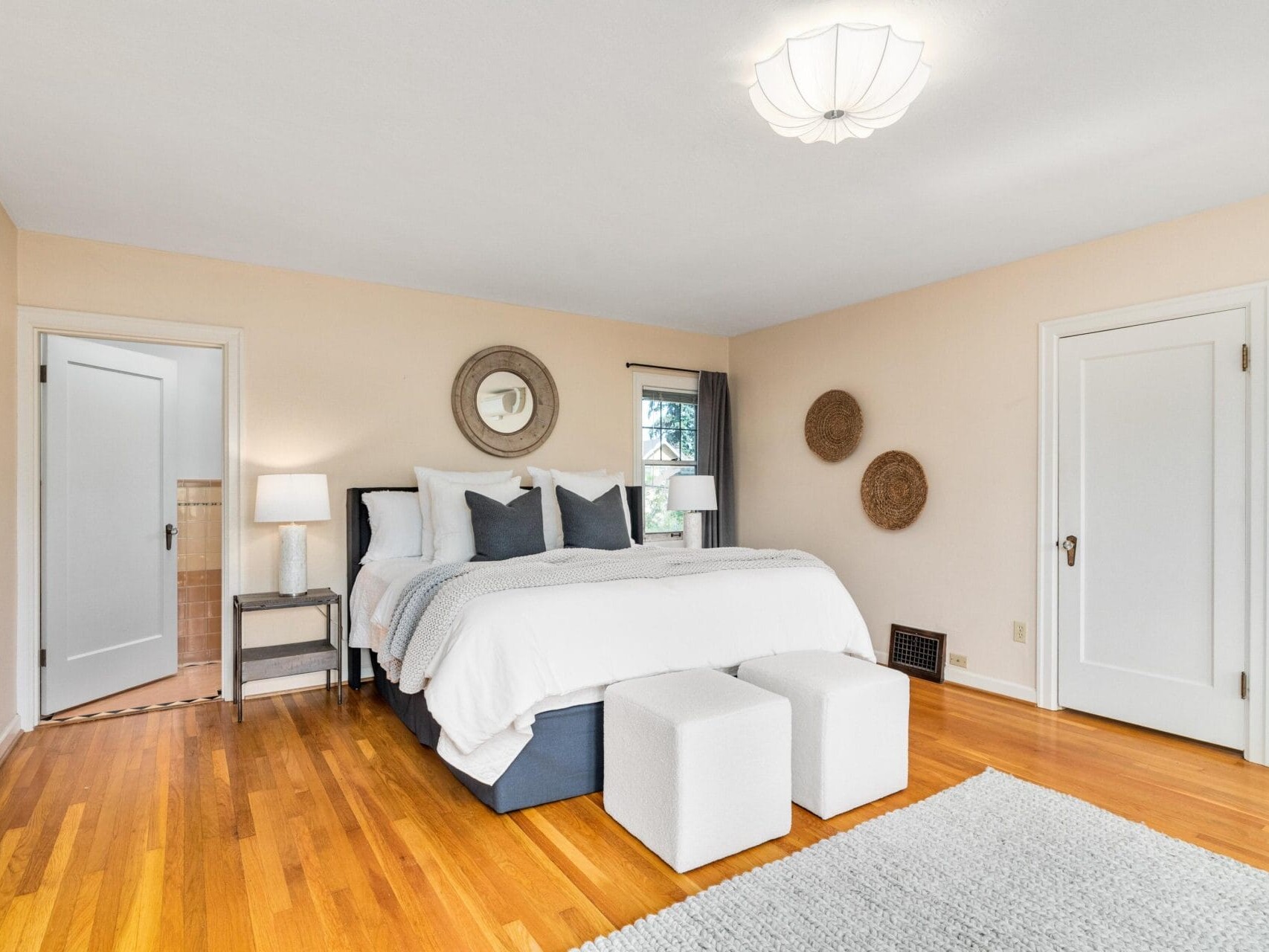 A bright bedroom with a large bed against a beige wall, adorned with gray and white pillows. Two white square ottomans are at the foot of the bed. Wooden floor, two closed doors, round mirror, and two wicker wall decorations are visible.