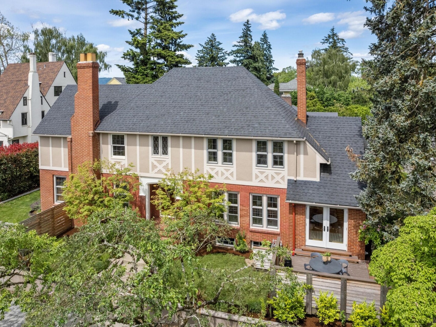 A large brick and timber two-story house with a dark roof, surrounded by lush greenery and trees. Theres a patio area with a small table and chairs. The home features several chimneys and windows, set in a suburban neighborhood.