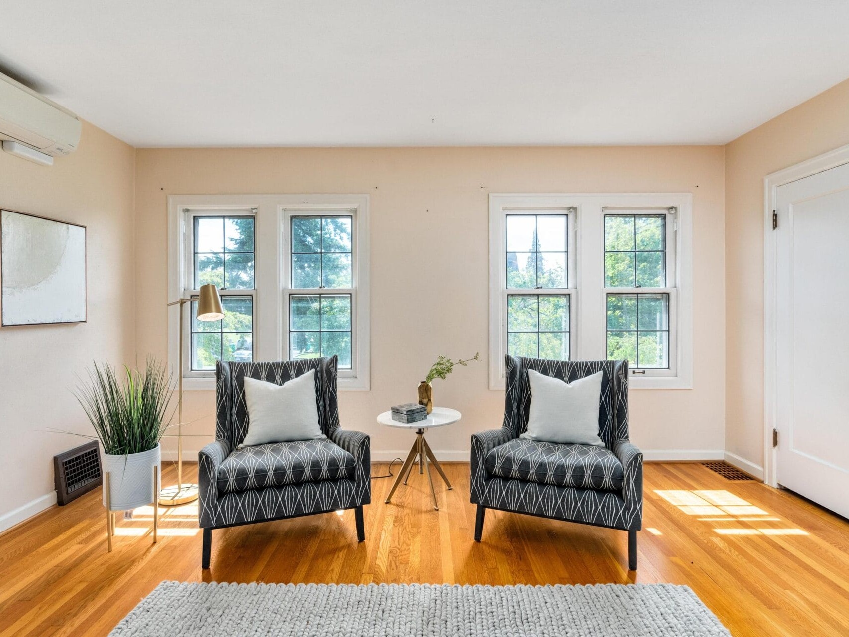 A bright living room with two patterned armchairs facing each other, a round white side table with a vase in between, two large windows, a floor lamp, and a rug on the wooden floor. The walls are light-colored with minimal decor.