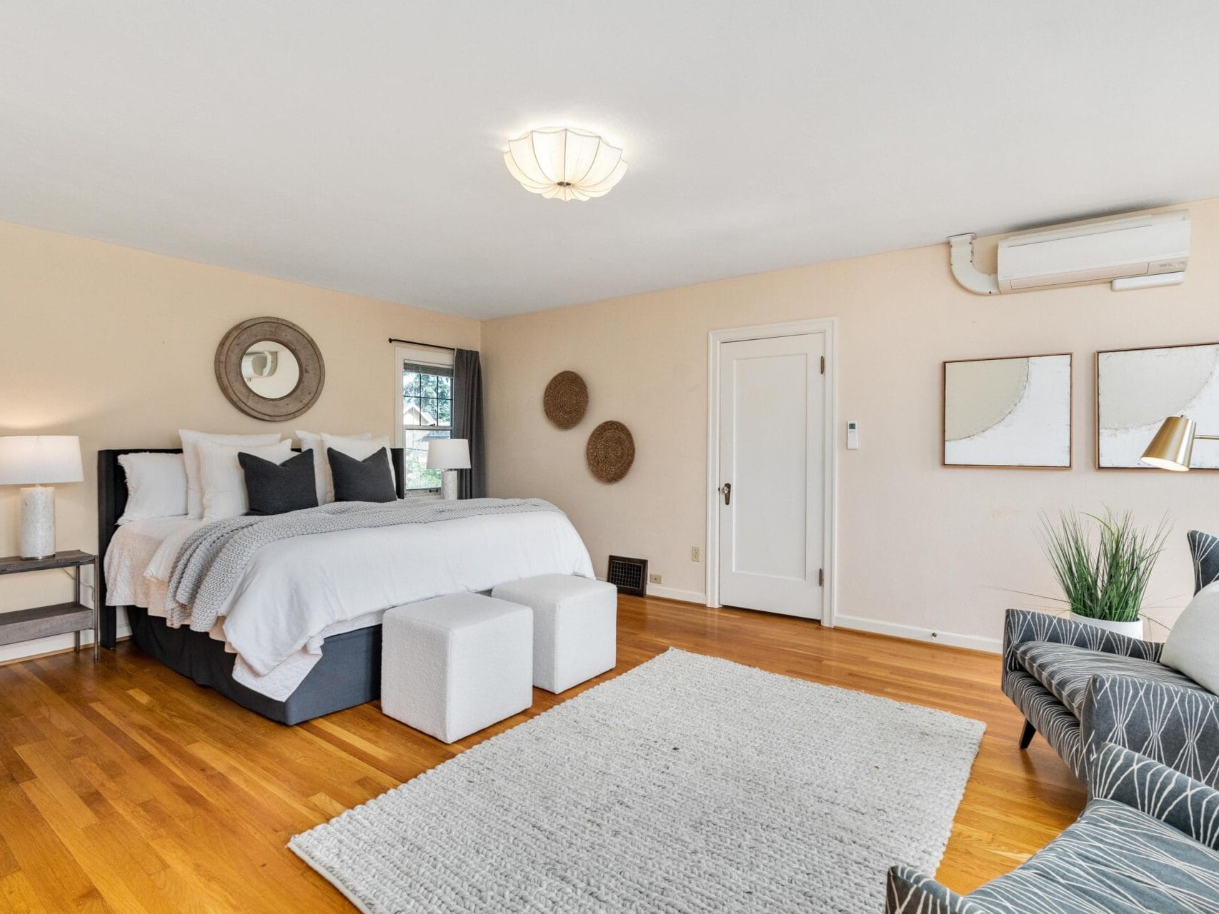 A spacious bedroom with a large bed adorned with white and gray bedding, two white ottomans at the foot, and a gray patterned chair. The room features hardwood floors, wall art, a potted plant, and a double lamp on a side table.