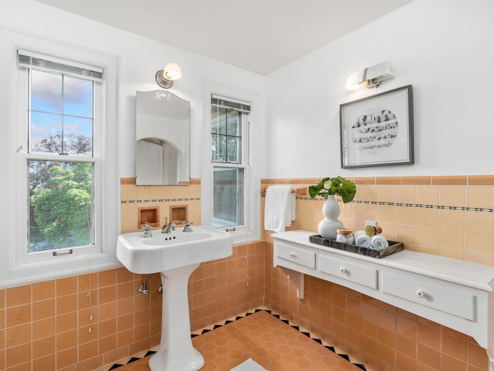 A bright bathroom with peach and white tiles features a pedestal sink, a framed mirror, and two windows. A wall-mounted light is above the mirror, and a white shelf holds rolled towels and a plant in a vase.