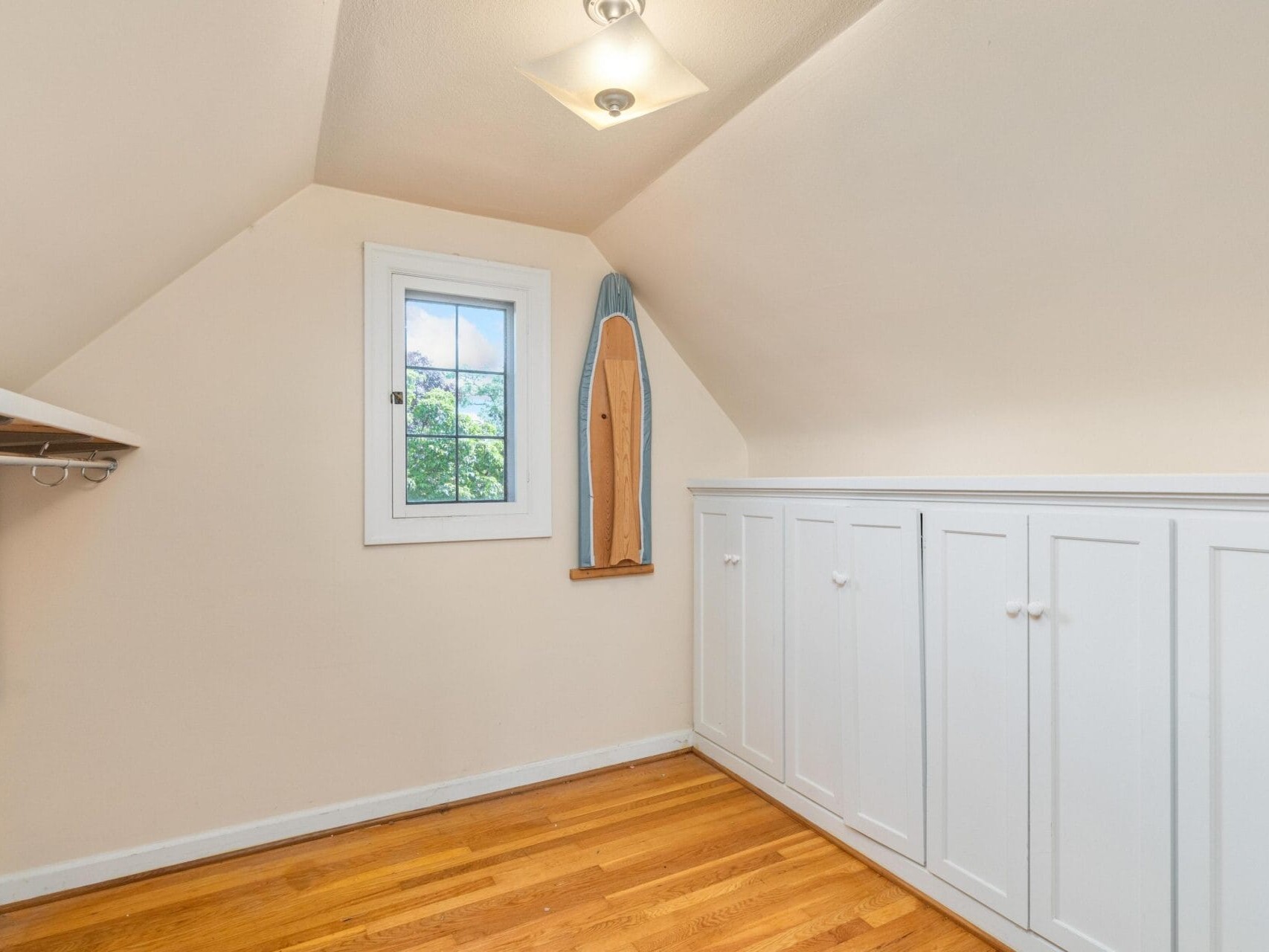 A small attic room with angled ceilings, featuring a single window and hardwood floors. A longboard is propped against the wall. White built-in cabinets line one side of the room with a wooden shelf on the opposite side.
