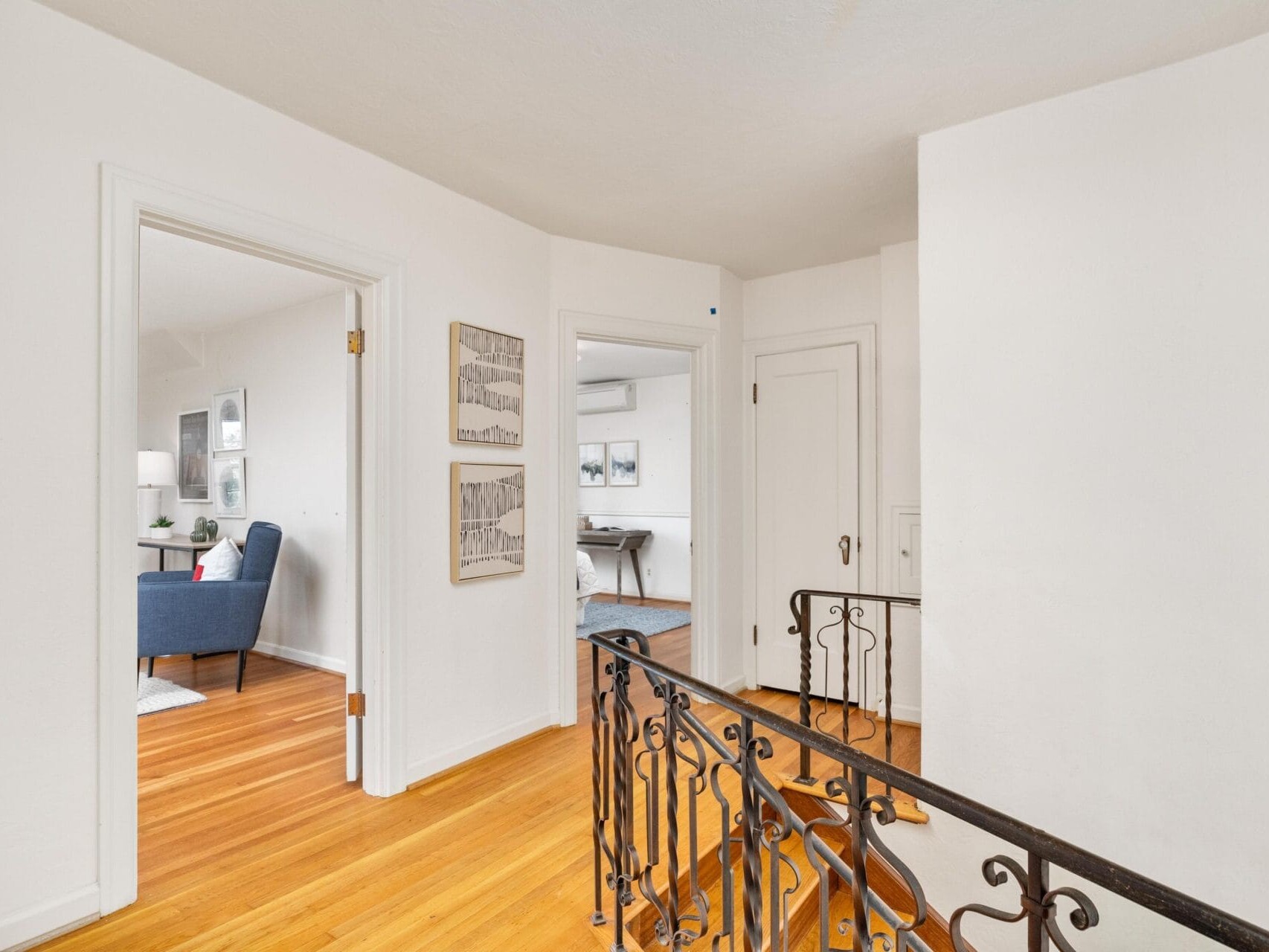 A hallway with light wooden floors and white walls leads to rooms. The foreground features a decorative iron railing. One room has a blue armchair and lamp, and theres wall art on the hallway walls. Natural light brightens the space.