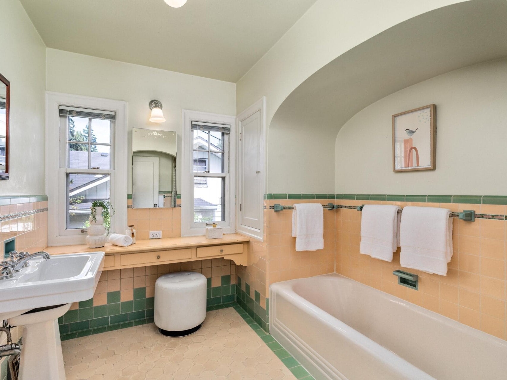 A vintage-style bathroom with light green and beige tiles, featuring a bathtub, a sink, a mirror, a window, and a small vanity table with a round stool. A towel rack above the tub holds three white towels. A framed picture hangs on the wall.