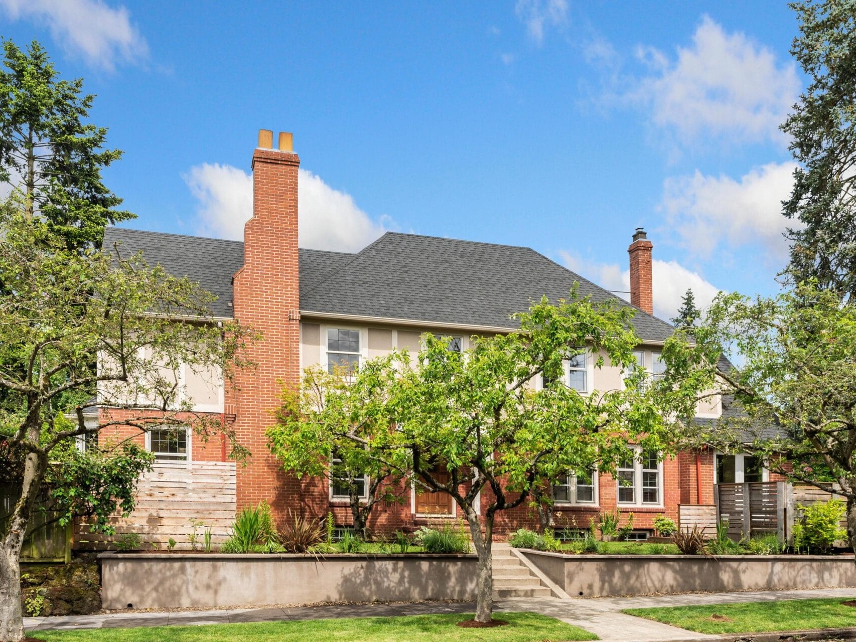 A two-story brick house with a dark gray roof, flanked by tall trees and surrounded by a manicured lawn. The sky is clear and blue, adding to the serene residential setting.