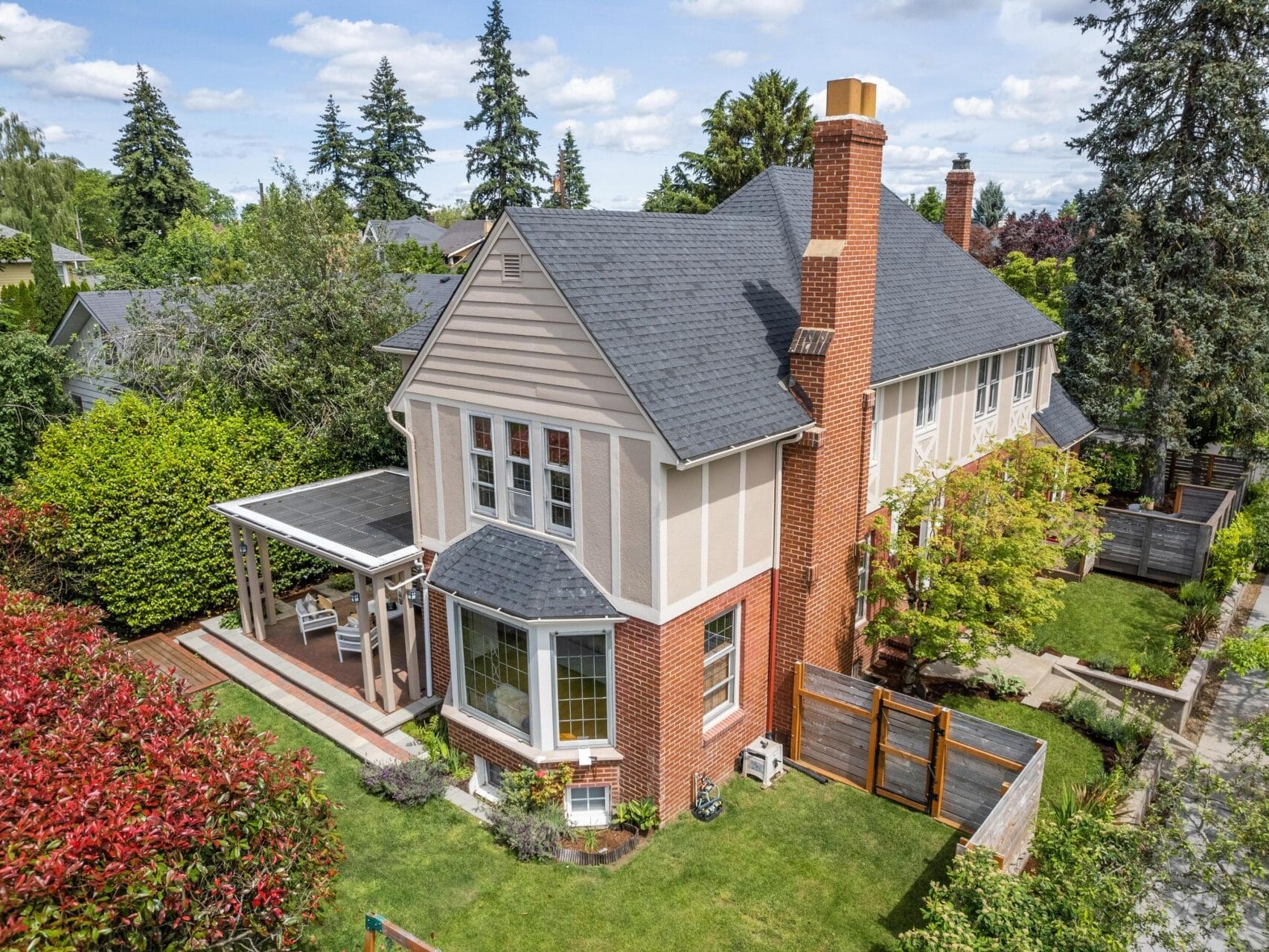Aerial view of a charming two-story house with a mix of brick and beige siding. It features a covered porch, bay windows, and a well-maintained garden. Tall trees surround the property, and a wooden fence borders the backyard.