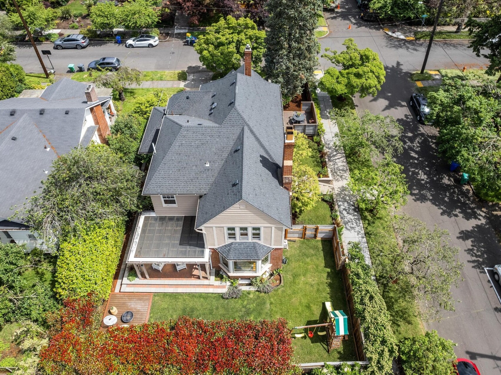 Aerial view of a two-story house with a gray roof, surrounded by greenery and shrubs. The backyard features a small lawn, playground equipment, and a patio. Neighboring homes and tree-lined streets are visible in the background.