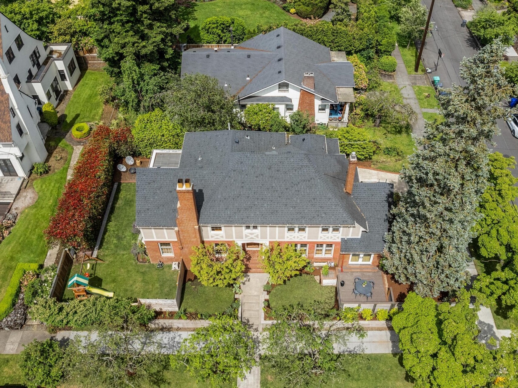 Aerial view of a suburban residential area featuring a large brick house with a dark grey roof surrounded by lush greenery. Other houses and a tree-lined street are visible. The garden includes a playset and patio.