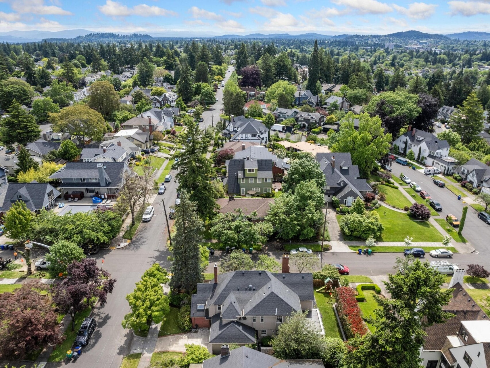 Aerial view of a suburban neighborhood with tree-lined streets and detached houses. The scene includes green lawns, neatly arranged homes, and distant hills under a partly cloudy sky.