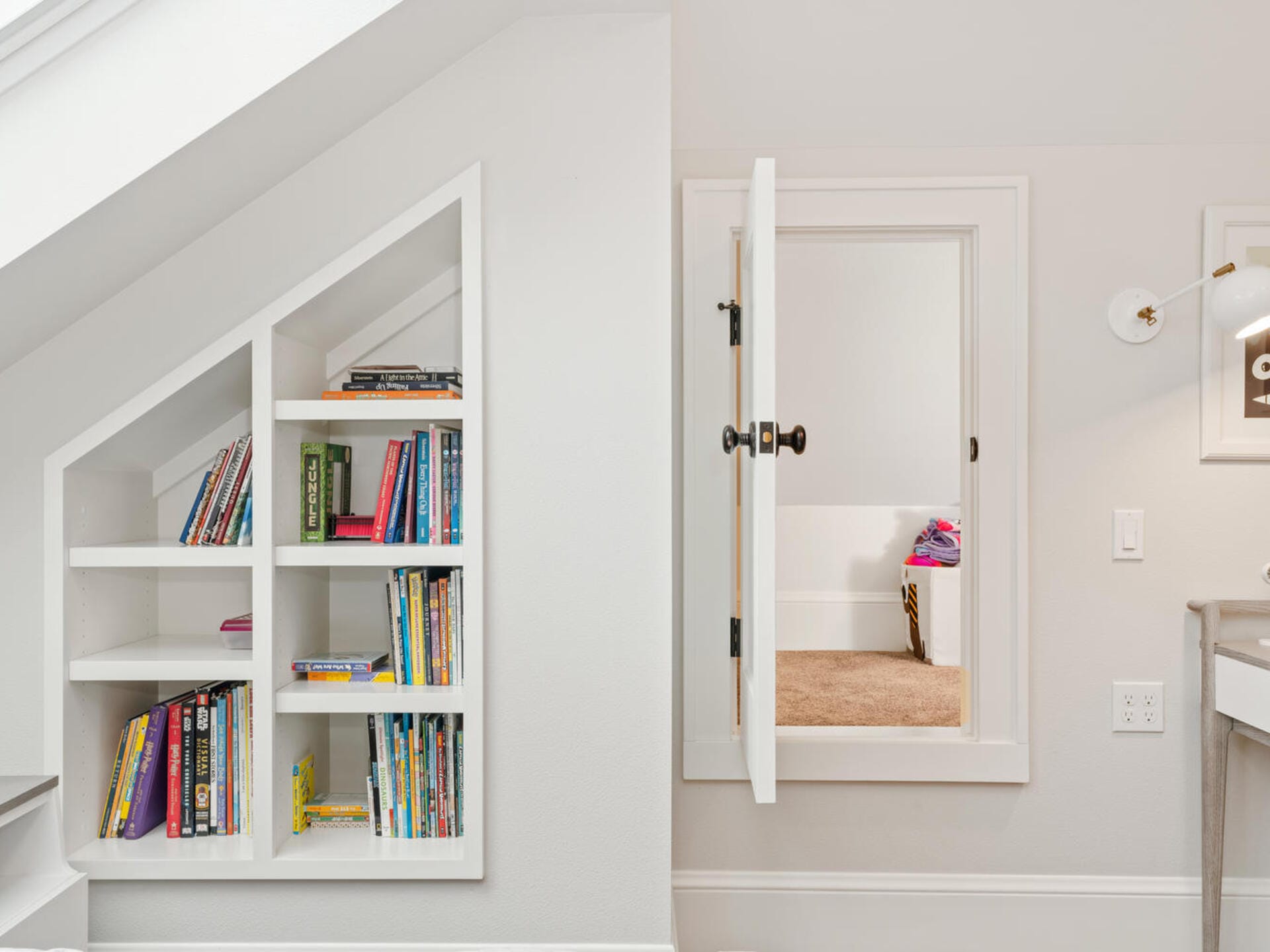 A cozy attic space features a unique built-in bookshelf with slanted shelves, filled with colorful books. To the right, an open door leads to a small carpeted room with a pink blanket visible inside. The walls and trim are painted white.