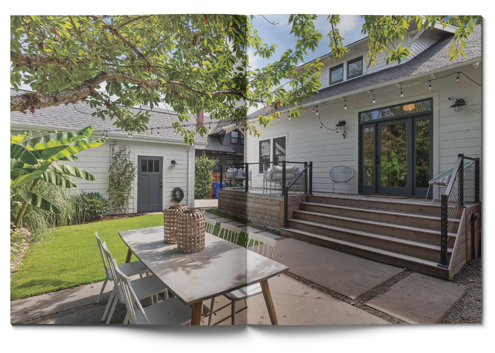 A backyard with a white house featuring a wooden deck, string lights, and steps. There is an outdoor dining table with chairs under a tree. The scene includes green grass, shrubs, and a small building with a door and window.