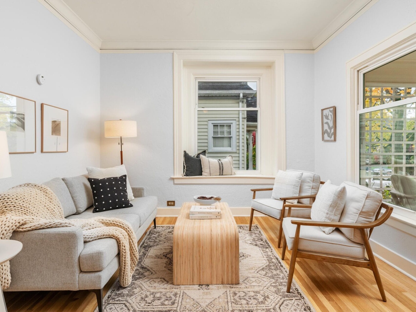 A cozy living room with a gray sofa and two wooden armchairs around a wooden coffee table. A lamp stands beside the sofa, and the room features a large window with an outdoor view. Decor includes framed wall art and a rug on hardwood flooring.