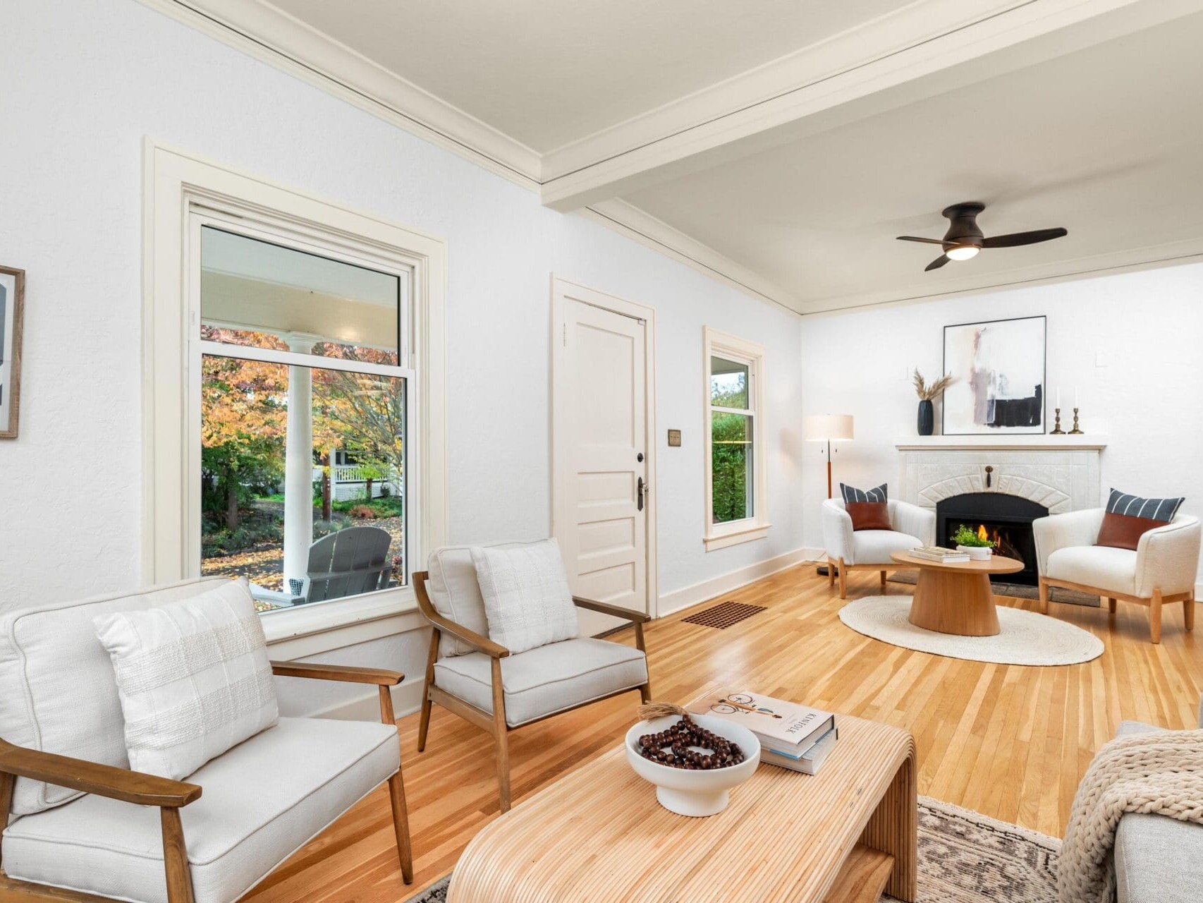 A cozy living room with wooden floors and light-colored walls. It features two armchairs, a sofa, a coffee table with a bowl of berries and books, and a fireplace. A large window shows colorful autumn trees outside. Theres a ceiling fan and artwork.