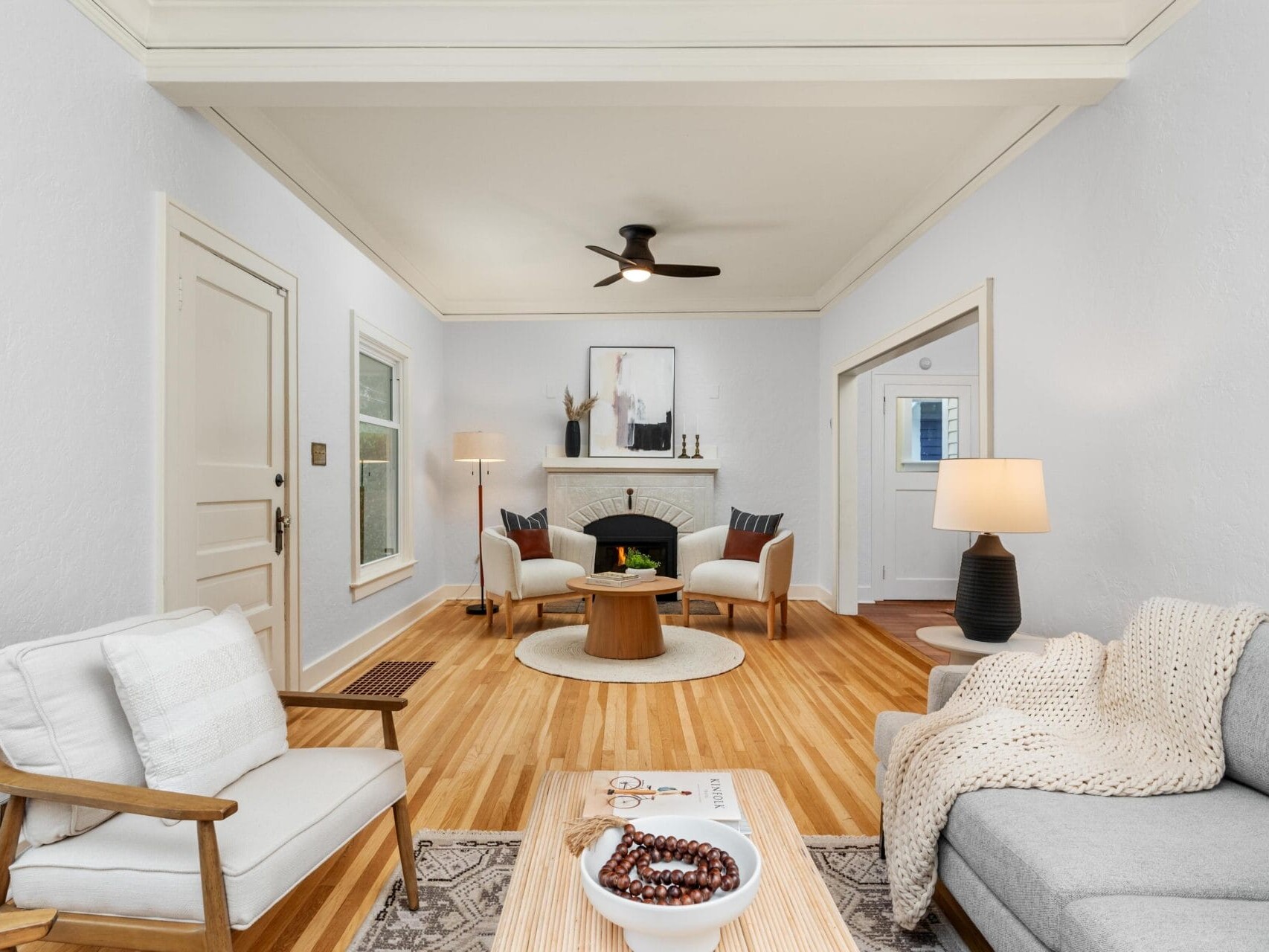 A cozy living room with light-colored walls and wooden flooring. It features a gray sofa, a wooden armchair, and a round coffee table. A ceiling fan is above, and a painting hangs above a decorative fireplace. Soft lighting creates a warm atmosphere.