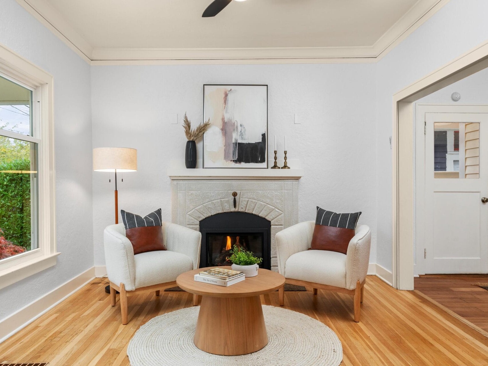 A cozy living room features a fireplace with a modern abstract painting above it. Two white armchairs with cushions flank a round wooden coffee table. A floor lamp stands to the left, and theres a doorway leading to another room on the right.