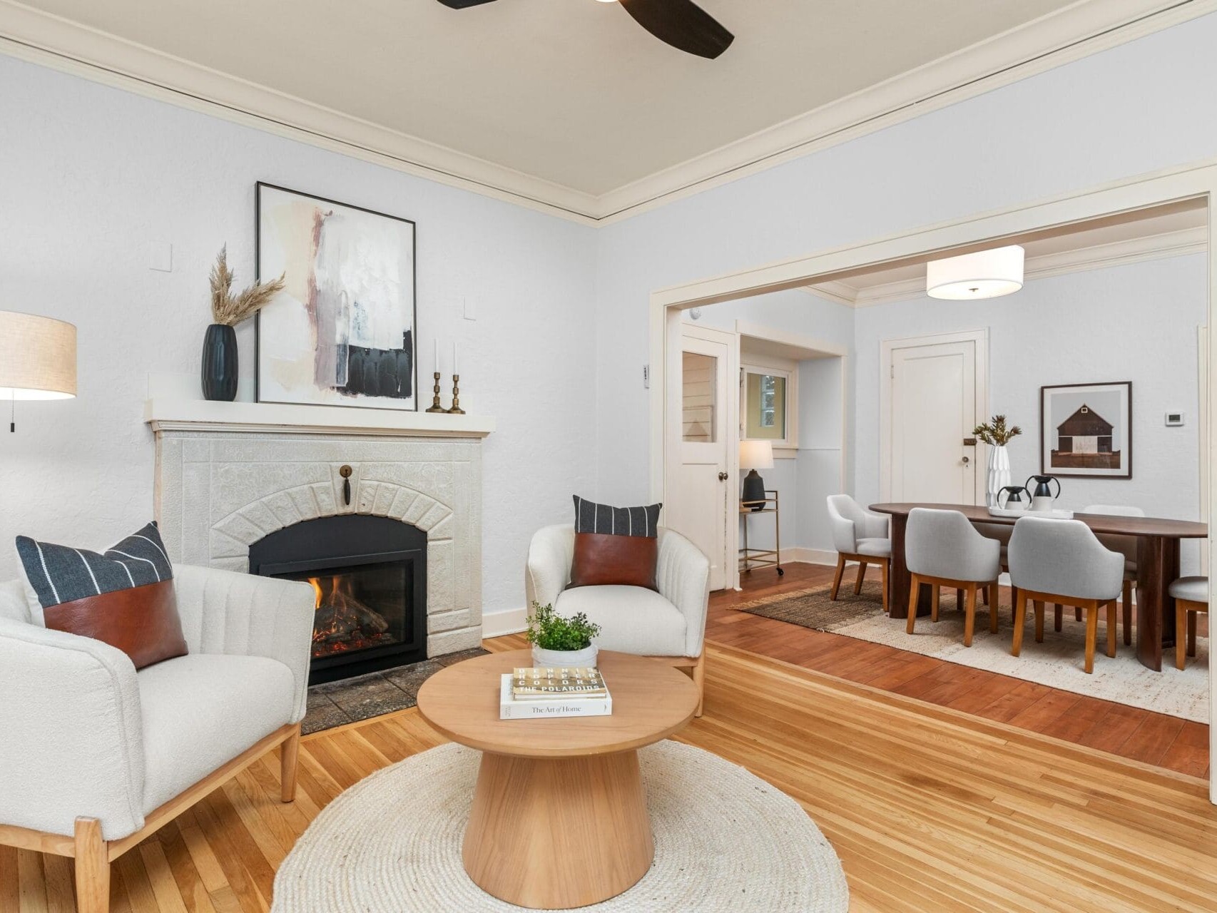 A cozy living room with a fireplace, two white armchairs, a round wooden coffee table, and a framed artwork above the mantel. The room leads to a dining area with a wooden table and chairs. Warm tones and hardwood flooring create an inviting atmosphere.