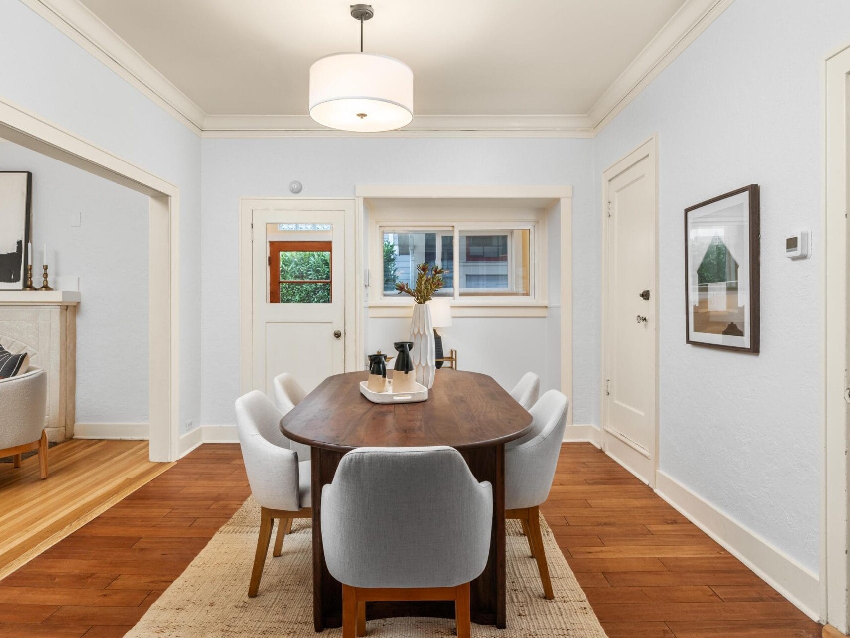 A dining room with a wooden table and six gray cushioned chairs on a light rug. Theres a modern light fixture above, a small window, and artwork on the walls. An open doorway leads to a living area with a fireplace.