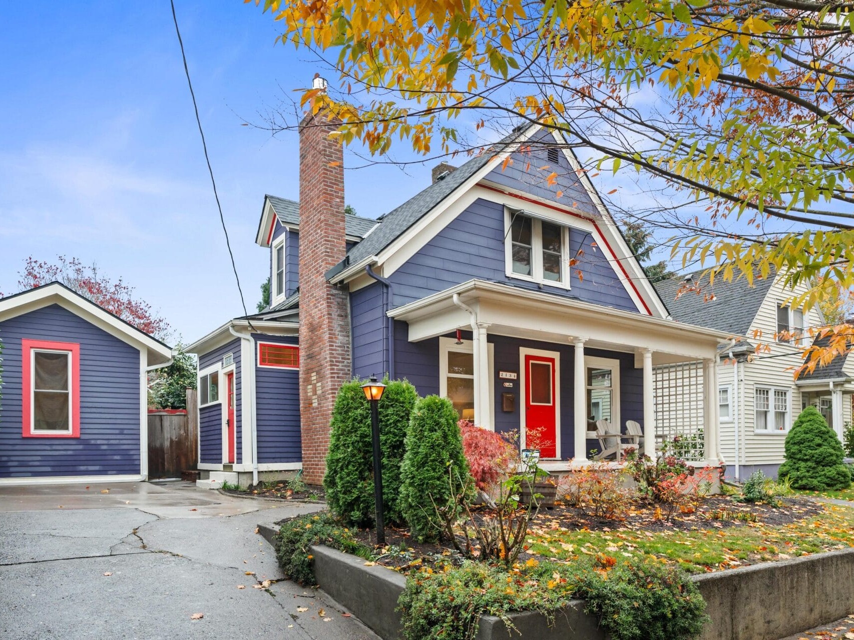 A charming blue house with white trim and a red door, surrounded by autumn foliage. A small detached garage is nearby. A paved driveway leads to the entrance, and a lush garden adds vibrant color to the front yard.