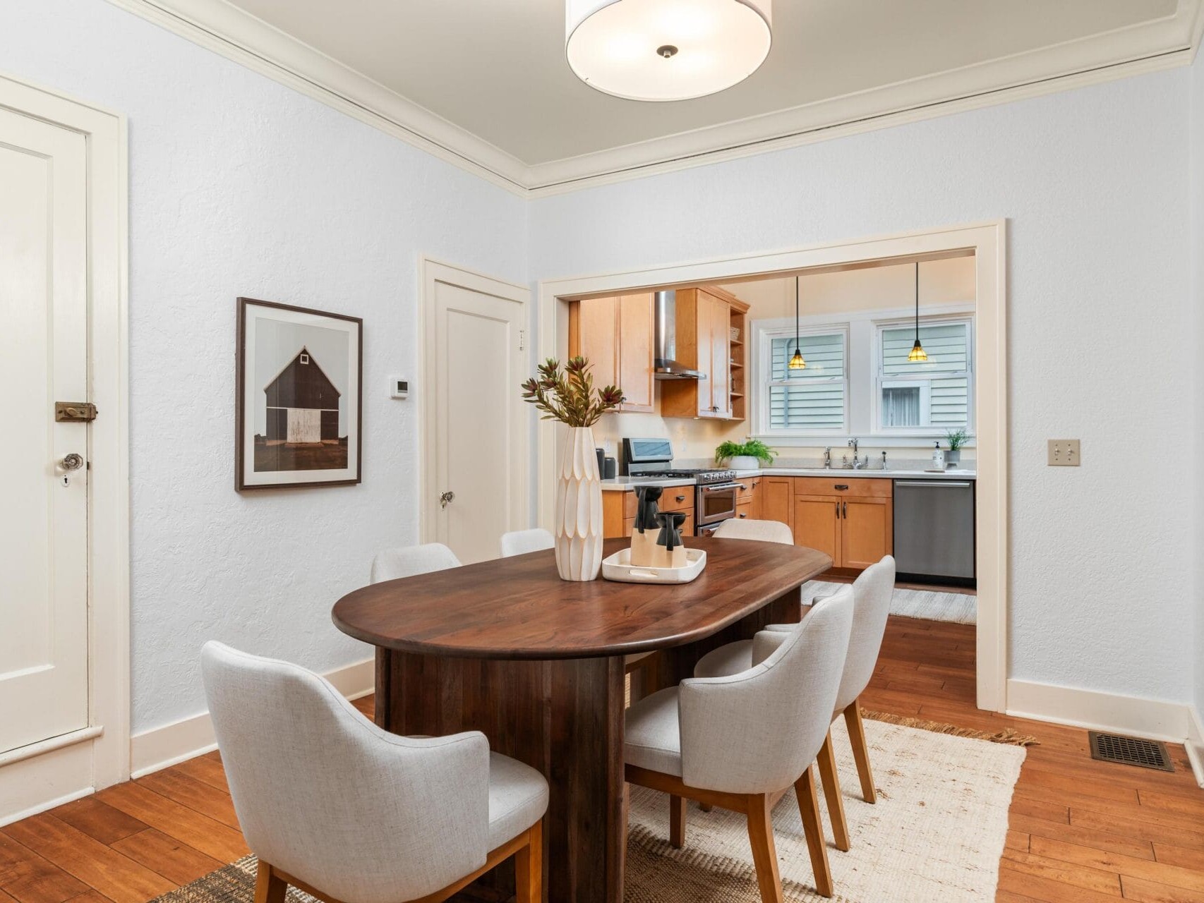 Dining room with a wooden table and six upholstered chairs. A modern vase with dried flowers is on the table. The kitchen is visible in the background, featuring wooden cabinets and a countertop with plants. White walls and wooden flooring complete the look.